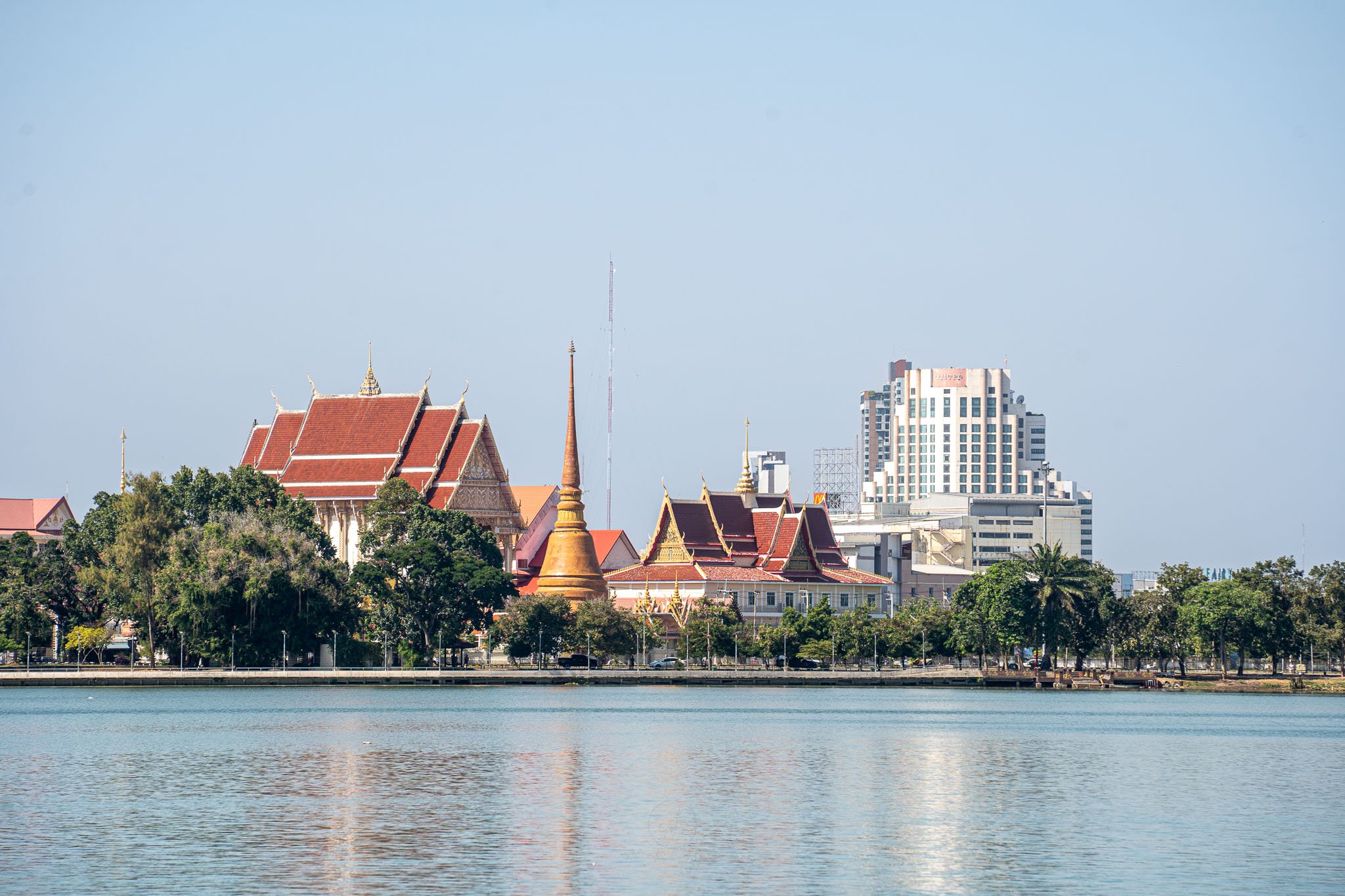 A view over the blue waters of Bueng Kaen Nakhon Reservoir. In the distance an orange stupa and prayer hall rise up to the blue sky.