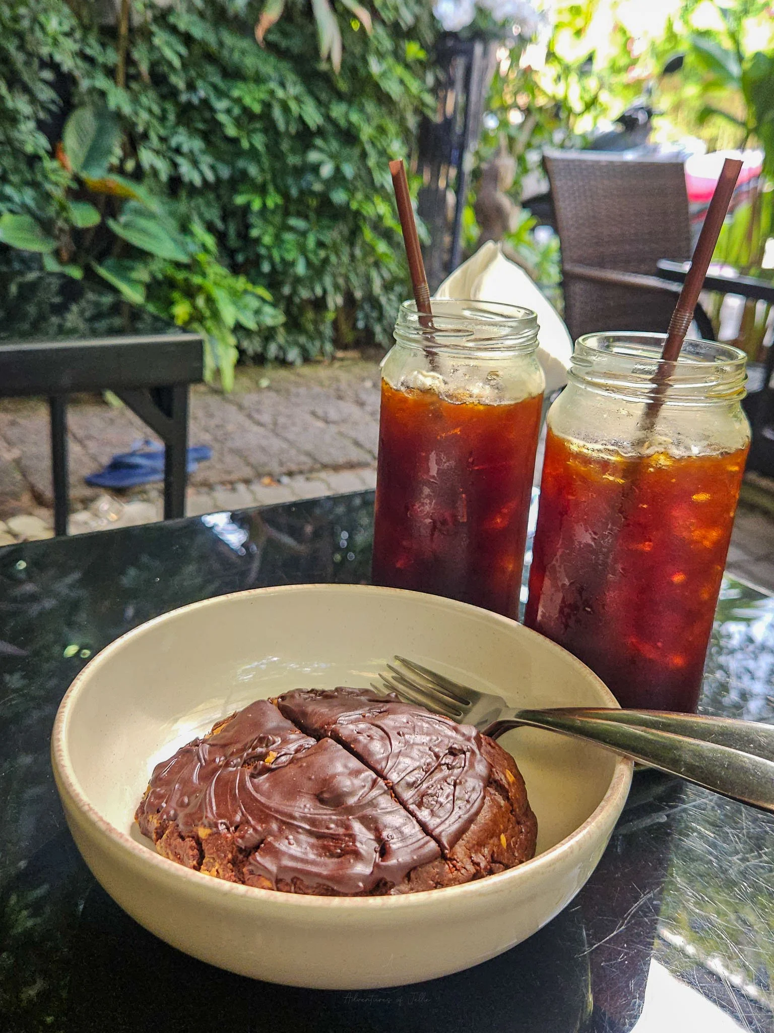 Sitting on a black glass table are two glasses of iced americano and a brown bowl filled with a chocolate covered peanut butter cookie. Photo taken in the garden of the Common Ground Cafe on Koh Phangan.