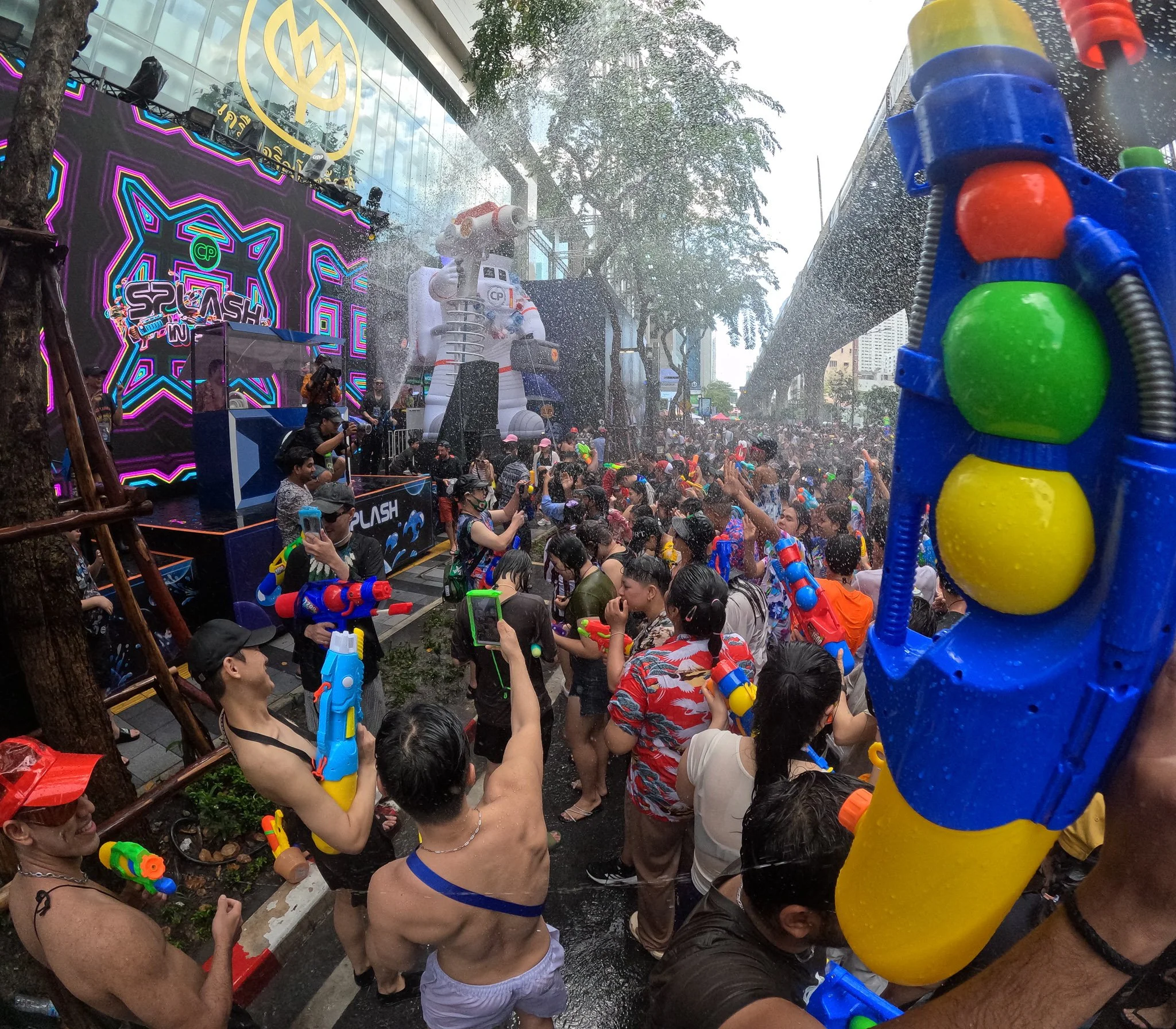 A large multi-colored watergun is raised. Behind it a Dj performs on a stage, the air looks like a rainstorm with all the waterguns being fired.