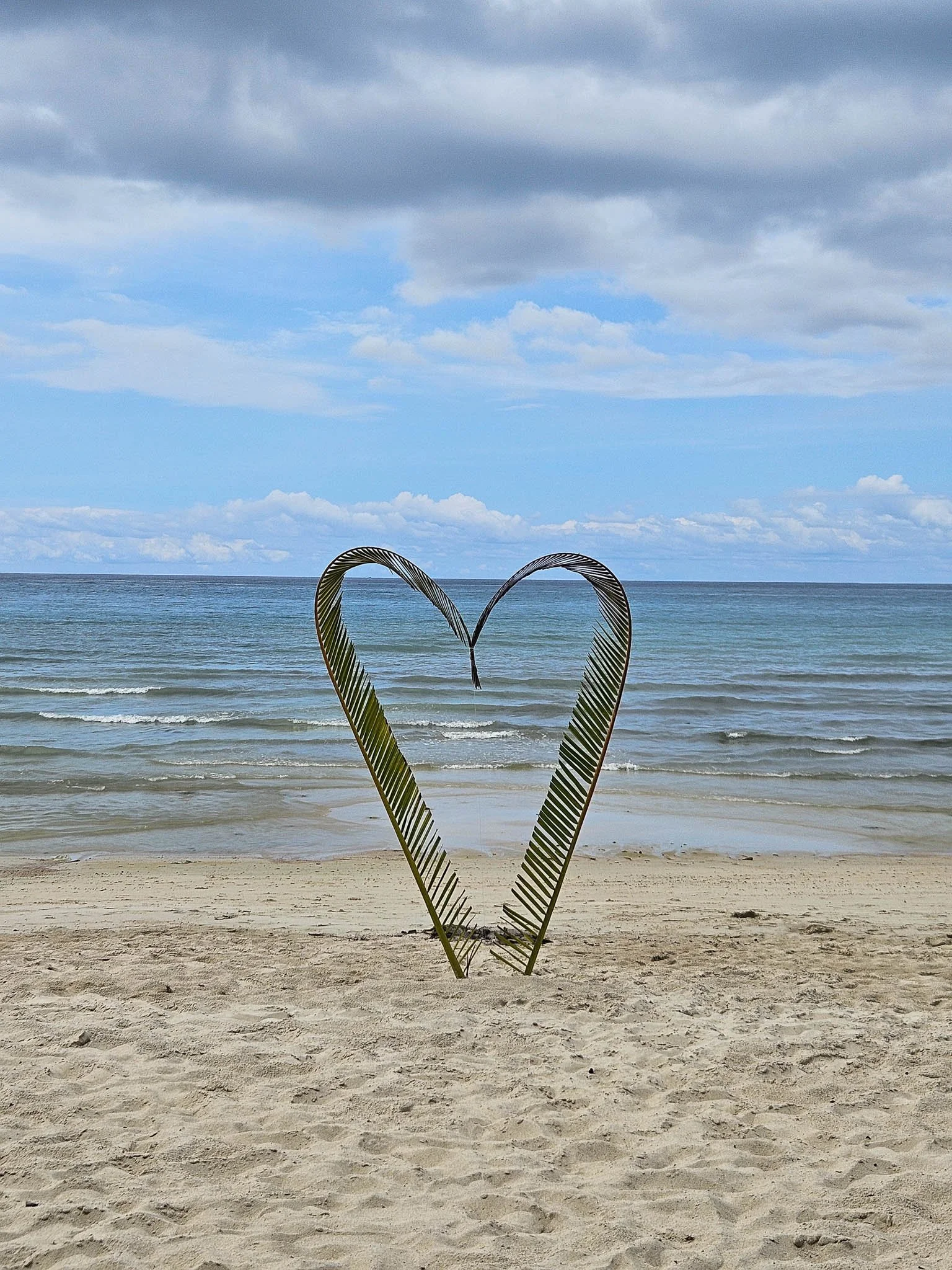 A heart made out palm leaves sits in the middle of the white sand on Bottle Beach, Koh Phangan. Behind, gentle waves lap at the shoreline.