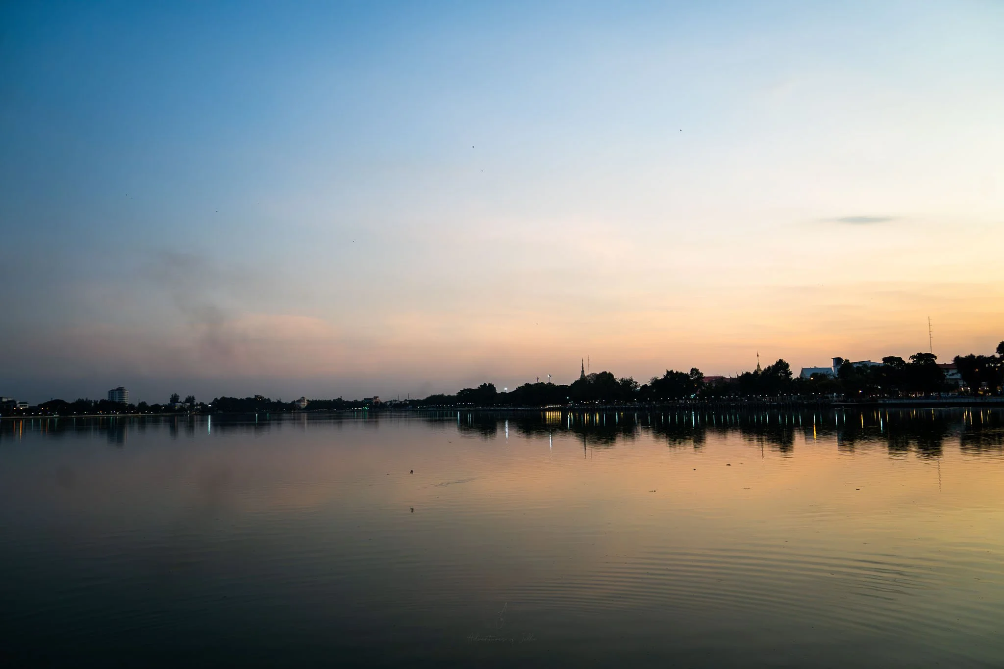 The sun sets over the Kaen Nakhon Lake. On the waters edge trees and buildings line the shore in silhouette.