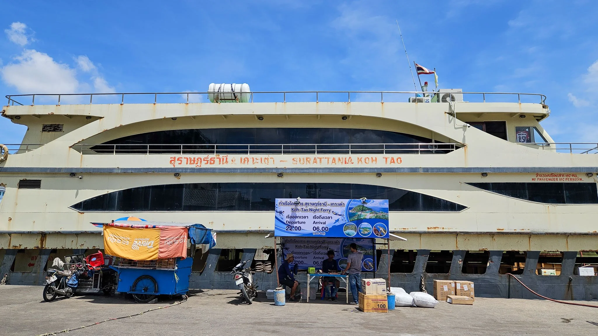 The Surat Thani to Koh Tao overnight ferry sits moored next to a riverside pier. In front is a ticket booth with a blue sign which shows the departure and arrival times.