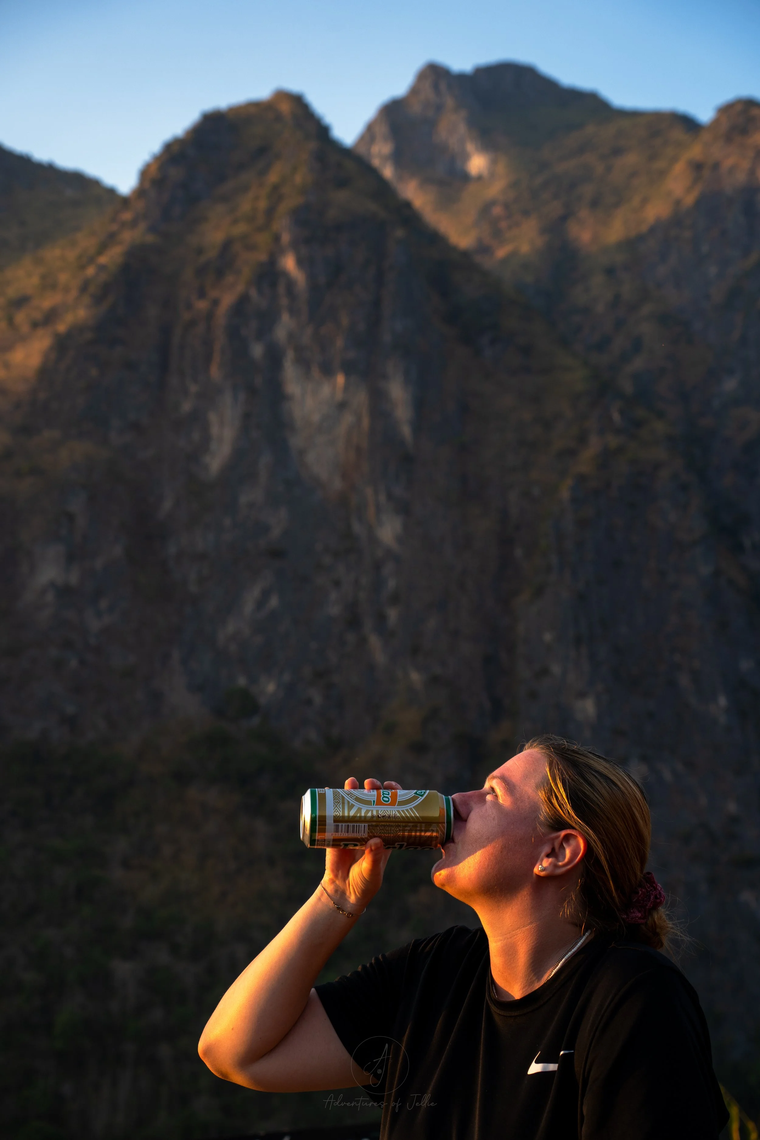Ellie looks up as she drinks a can of Beer Laos, her face bathed in golden light from the setting sun. Behind rocky mountains rise up to the blue sky above.