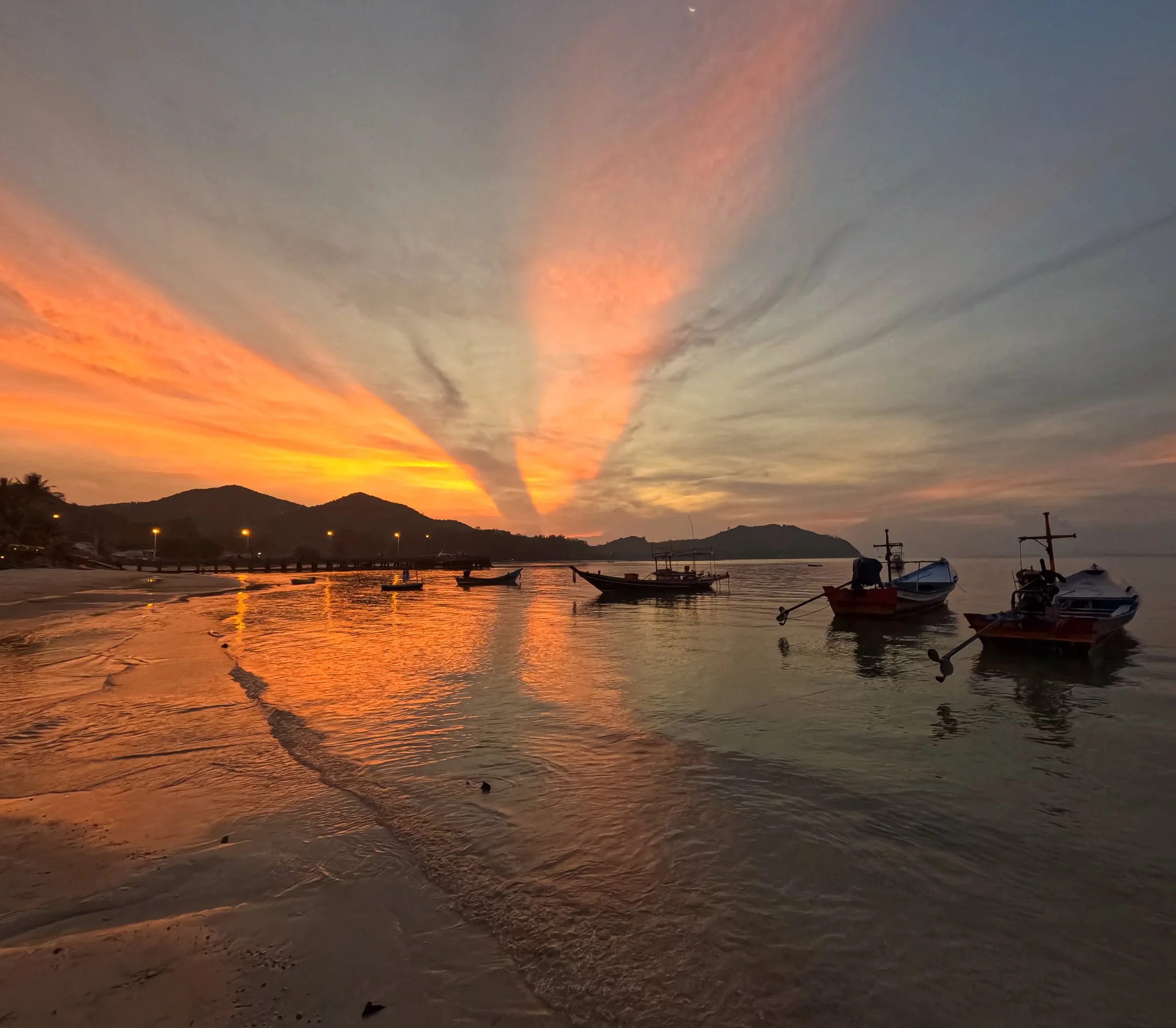 Longtail boats sit moored on the shoreline on Chaloklum Beach. Above in the sky, the setting sun is creating a fiery orange, pink and yellow sky.
