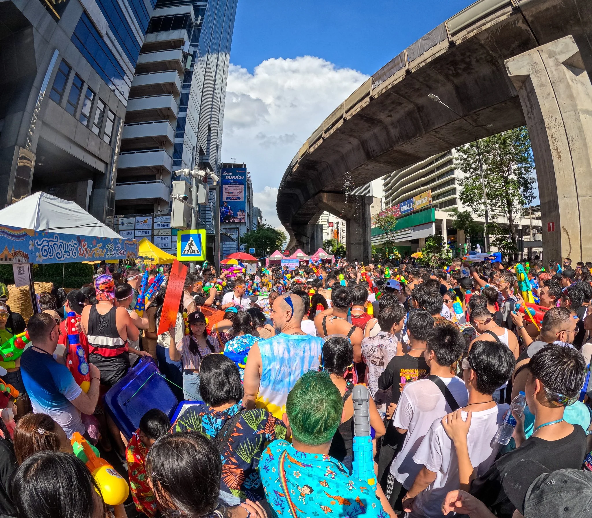 A blue sunny sky shines down on the Silom Songkran party goers. Jets of water arc from the crown and a cloud looms in the distance.
