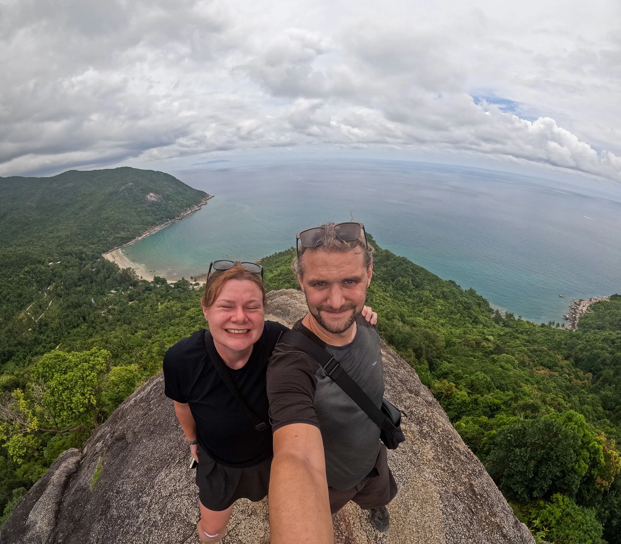 Adventures of Jellie stand at the edge of a rock overhanging jungle and the steep coastline below. Jellie pose for a selfie wearing black sportswear and sunglasses.