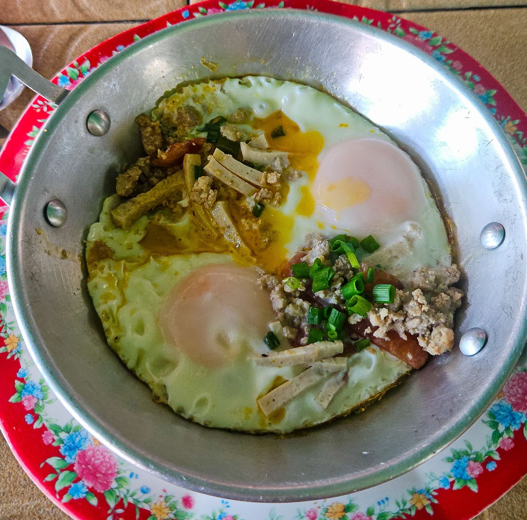 Two fried eggs sit in a metal dish topped with strips of Vietnamese sausage, sliced spring onion and Chinese sausage.