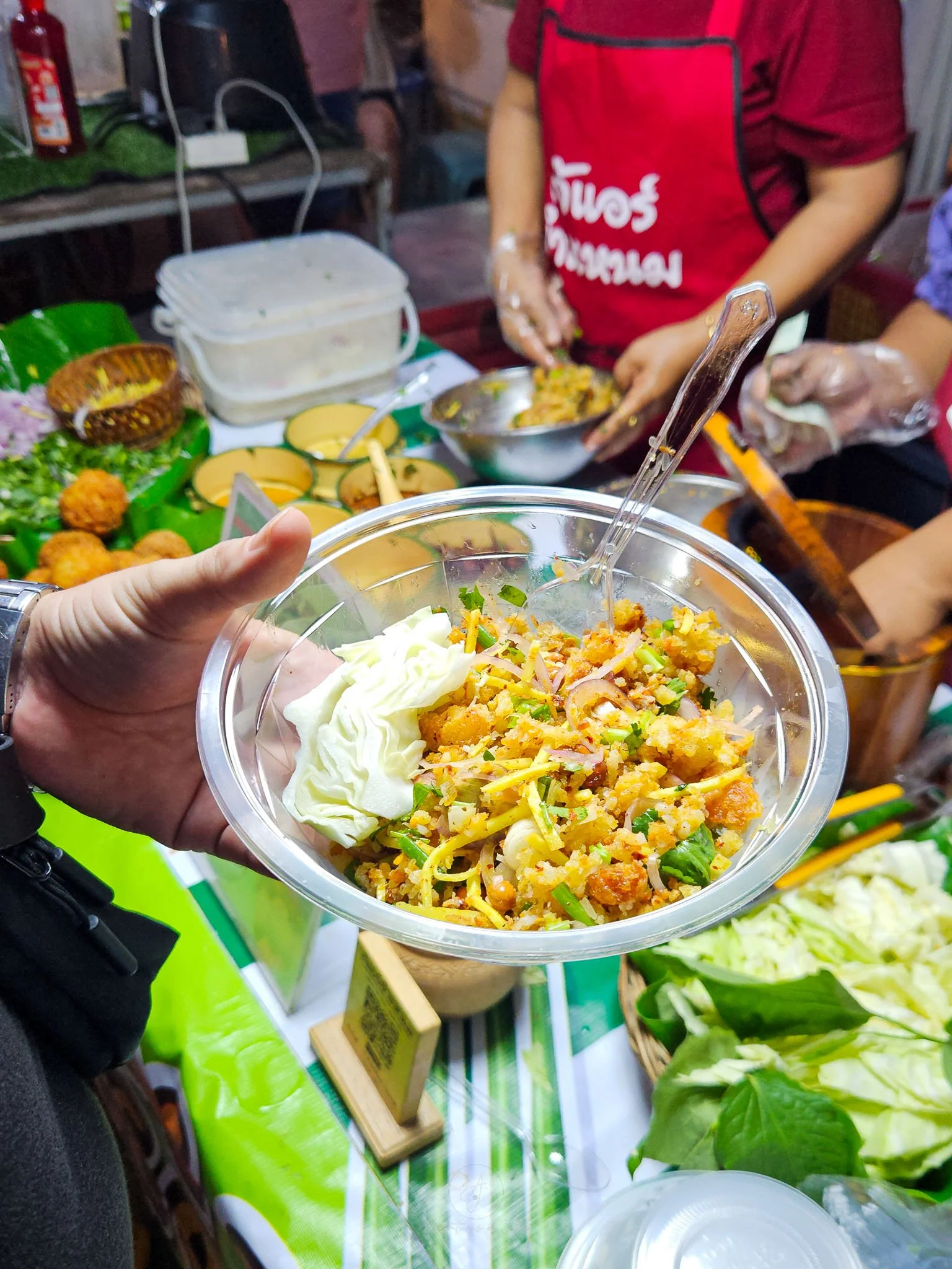 A bowl of yam naem khao tod (crispy rice salad with slices of red onion, peanuts and bamboo shoots) is held up to the camera in front of a market stall at the Chaloklum Sunday Night Walking Street Market.