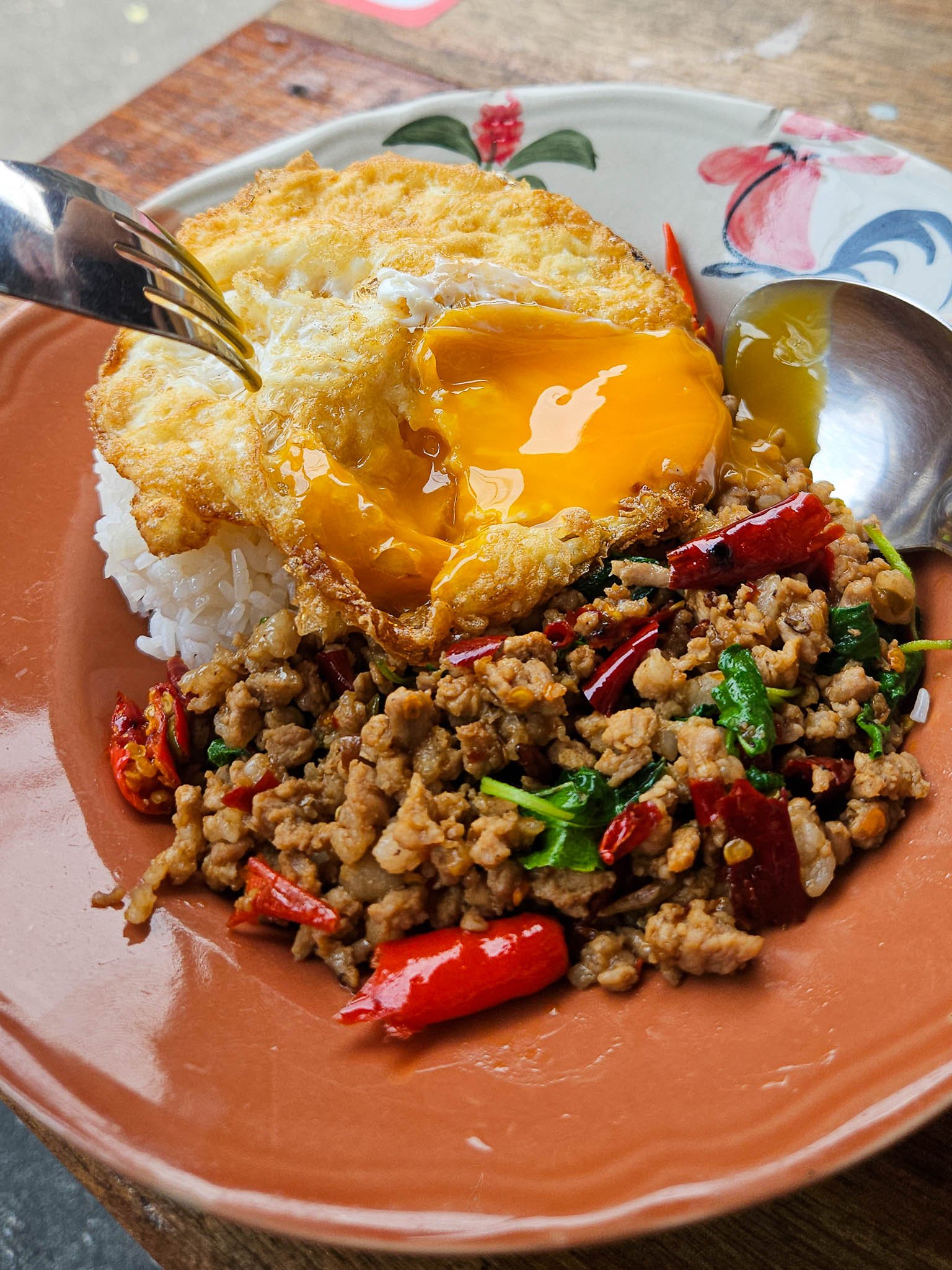 A fork breaks the yolk of a bright yellow, Thai style fried egg. The egg is sitting on top of a plate of pork pad kra pao studded with red chillies and green basil leaves.