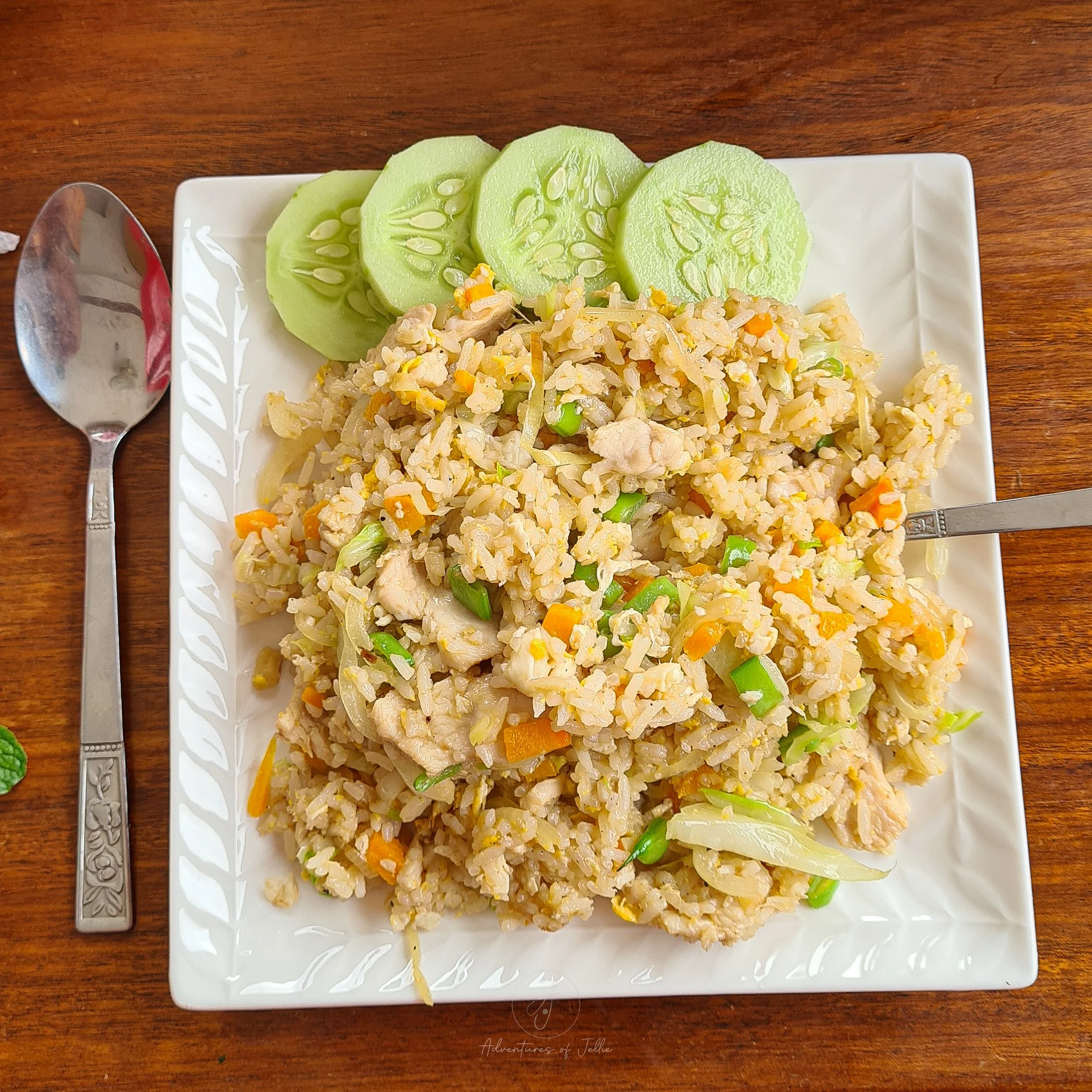 A white square plate is topped with a pile of chicken and vegetable fried rice and four slices of cucumber at the Namlin Restaurant in Nong Khiaw, Laos.
