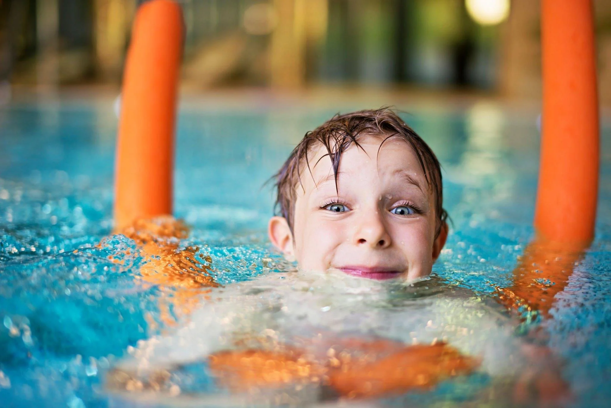 A young boy with wet hair smiling in a swimming pool, holding onto orange pool noodles.