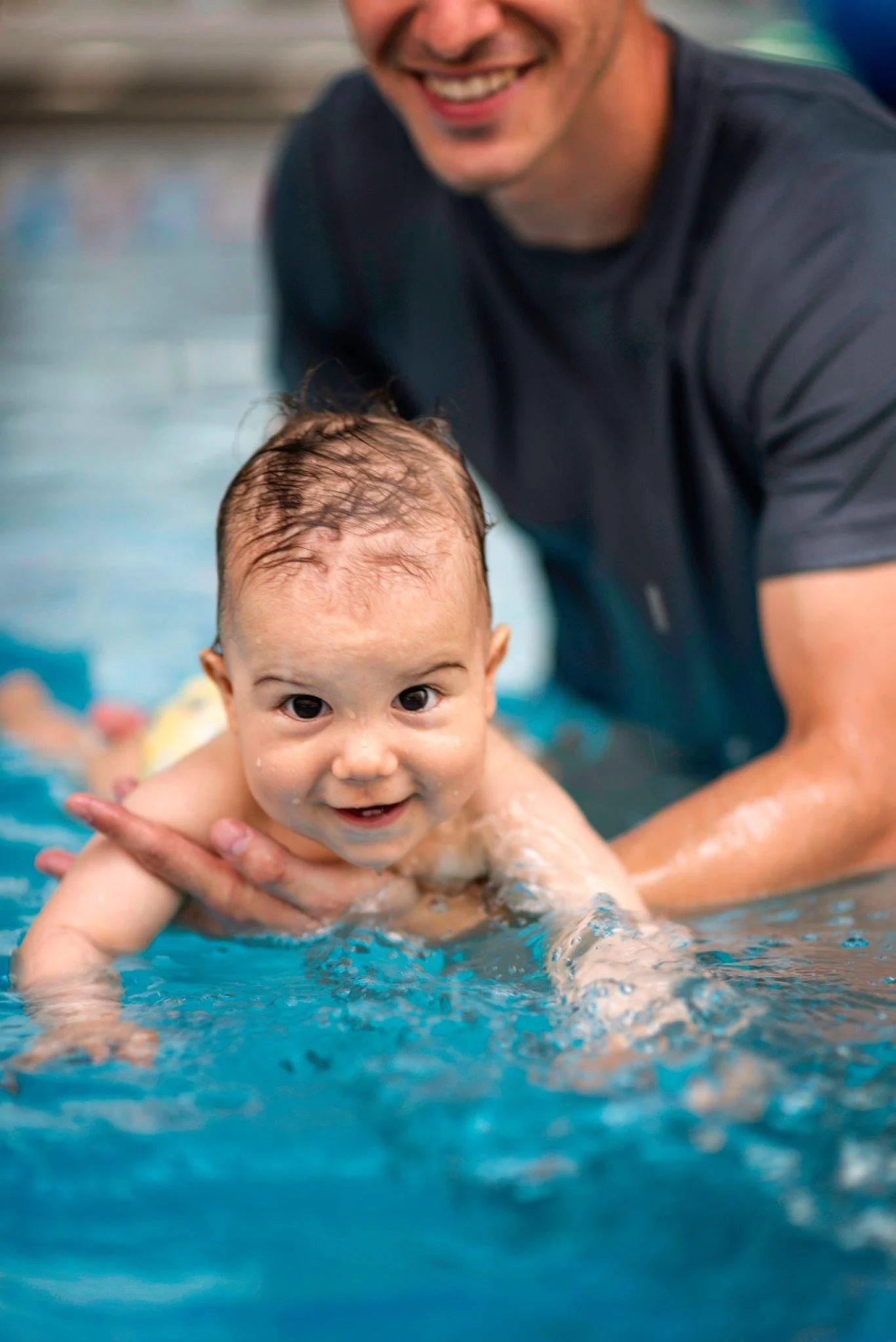 A smiling adult man holding a young child in a swimming pool, with the child looking at the camera.