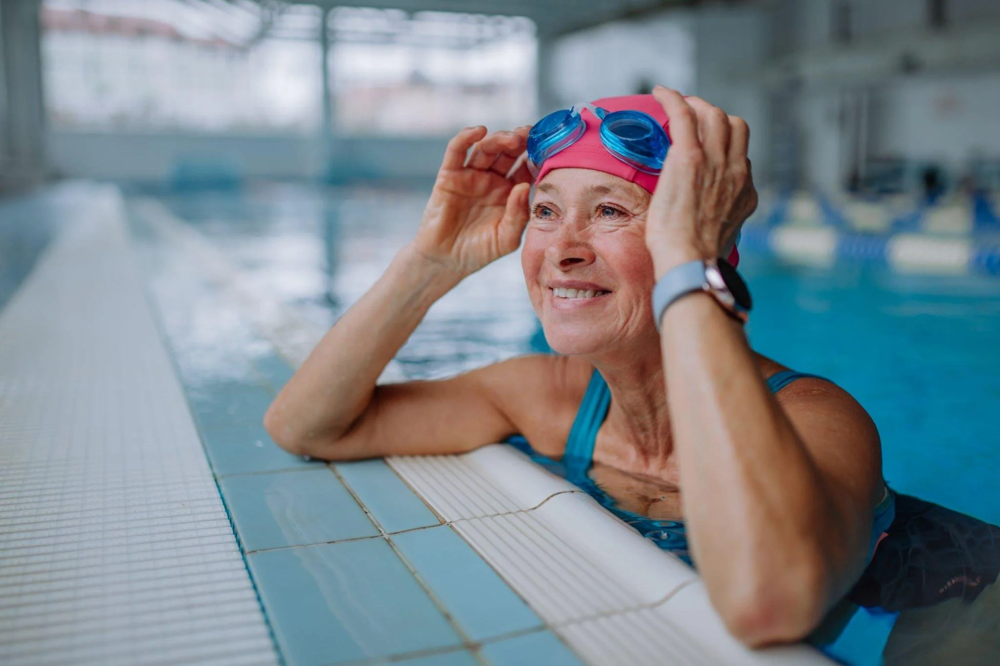 An elderly woman in a swim cap and goggles smiling while holding her goggles on her head, sitting at the edge of an indoor swimming pool.