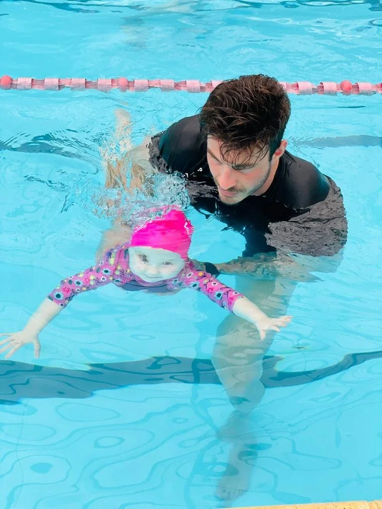 A man helps a young girl learn to swim in a swimming pool. The girl is wearing a pink swim cap and a colorful long-sleeve swimsuit. The man is underwater, supporting the girl as she practices swimming.