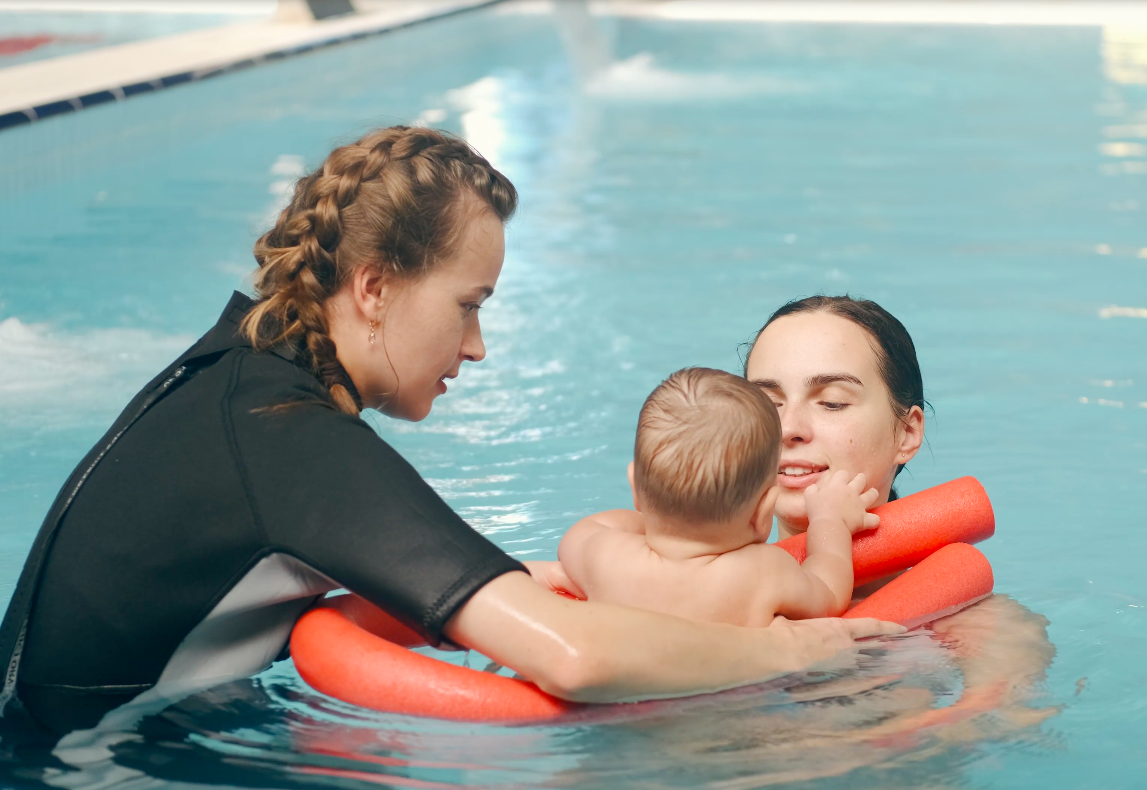 A woman holding a baby in a swimming pool with the assistance of another woman, all three are engaging in a swimming lesson.