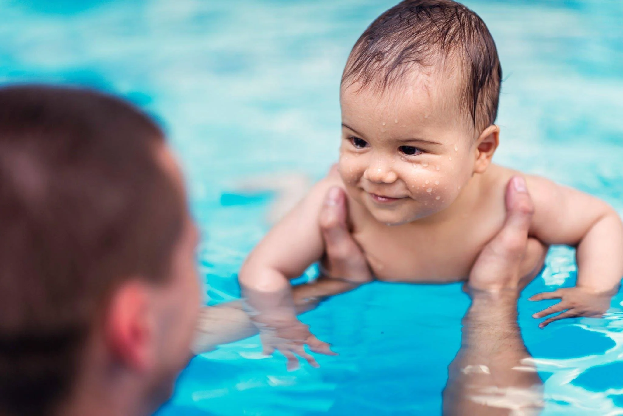 A smiling baby with wet hair being held in a swimming pool by an adult, with the baby's face and shoulders above the water.