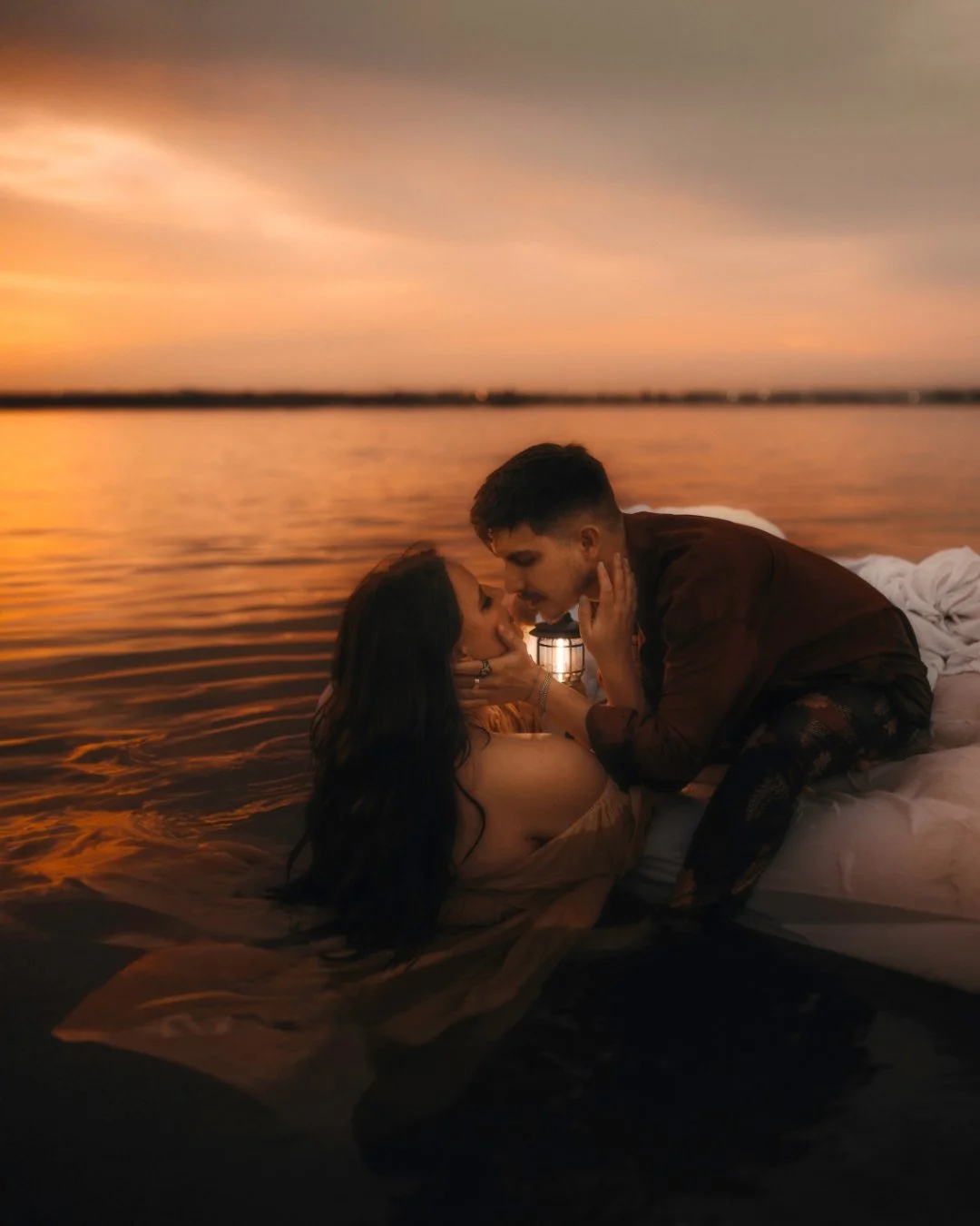 A romantic scene with a couple lying on a bed by the water during sunset, facing each other closely and touching faces, with a small lantern between them on a small boat.