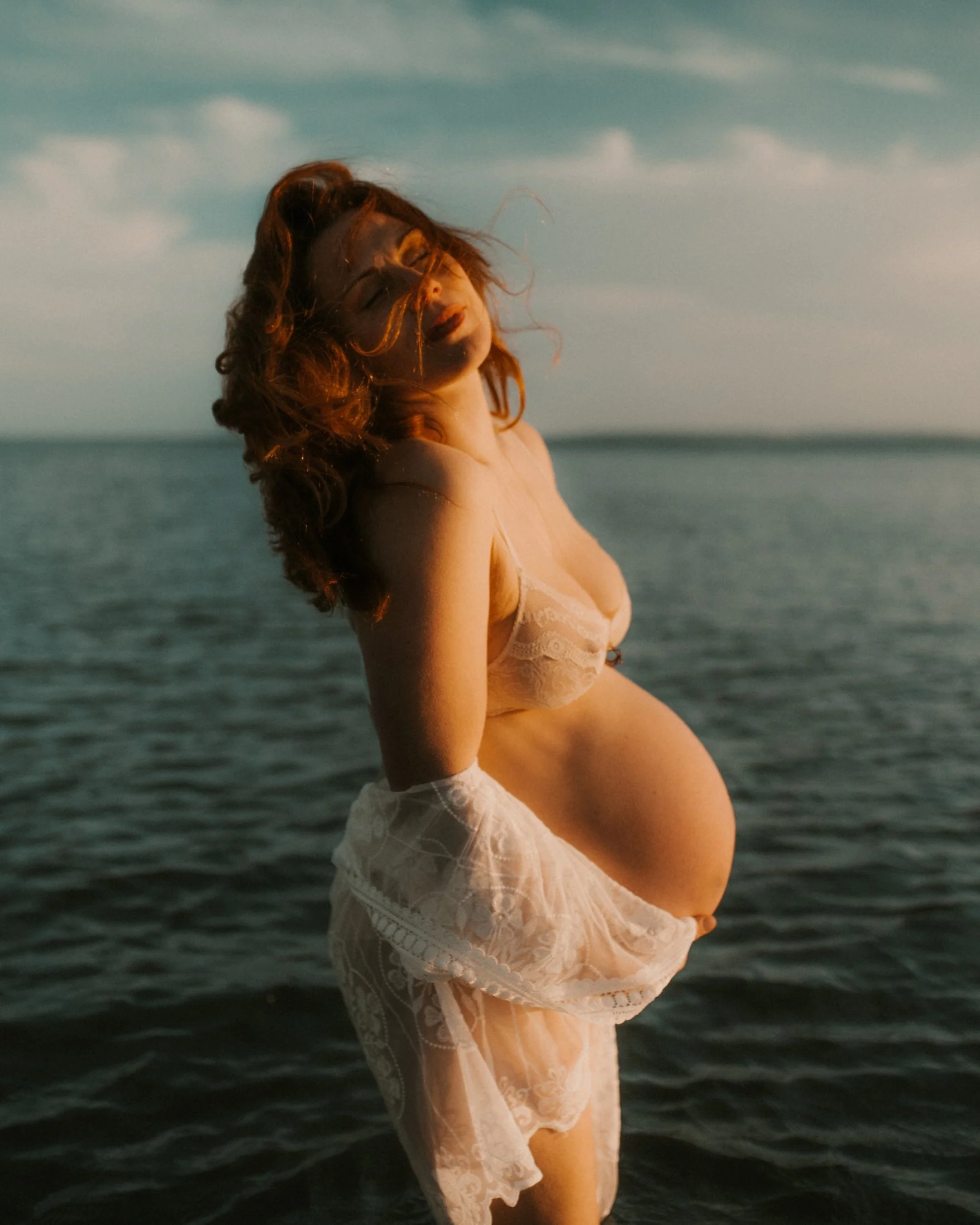 A pregnant woman with red hair stands in front of a body of water, wearing light-colored lacy lingerie and a matching skirt, with the sky and water in the background.