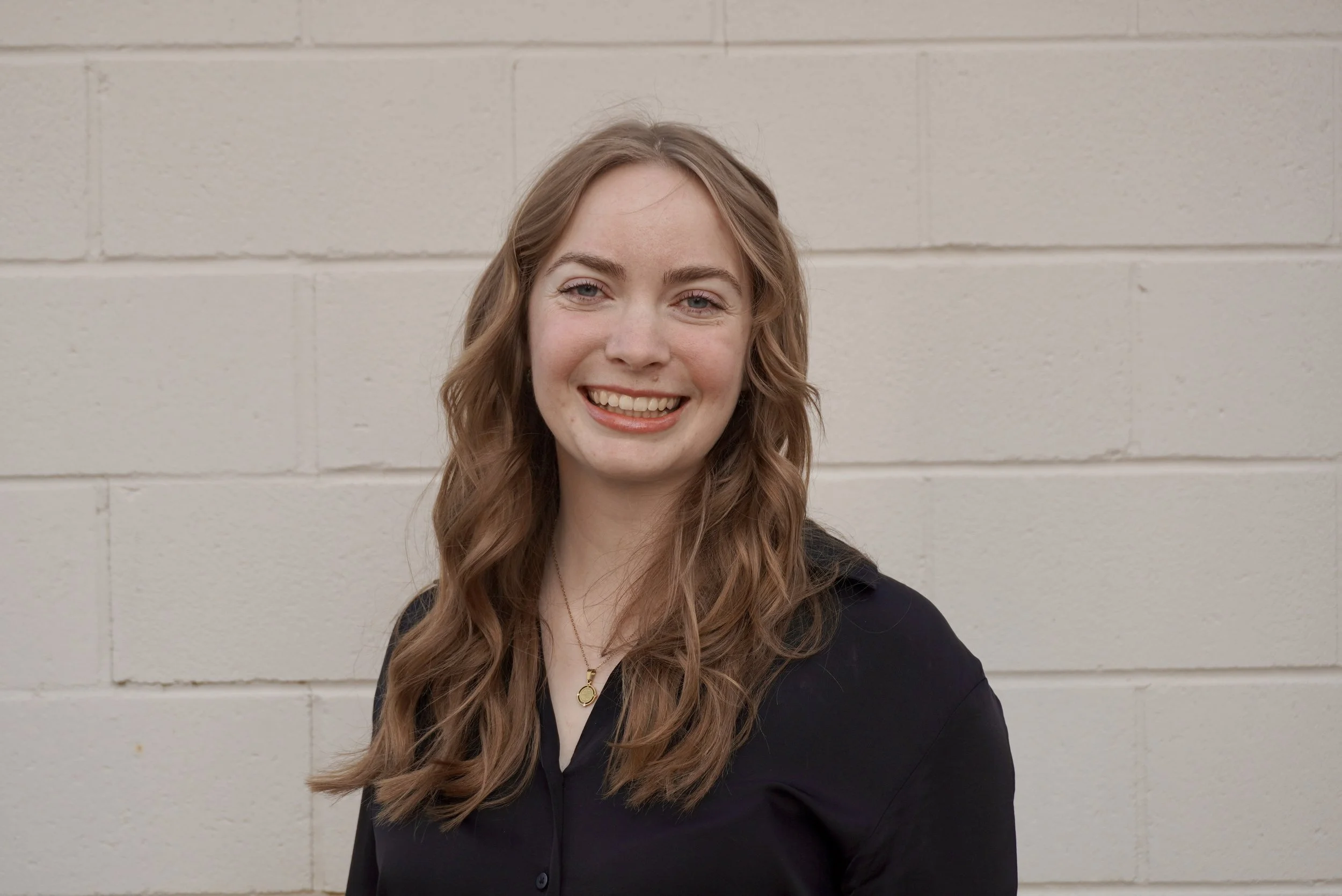 A young woman with long, wavy red hair and a bright smile stands in front of a beige brick wall, wearing a black shirt and a gold necklace.