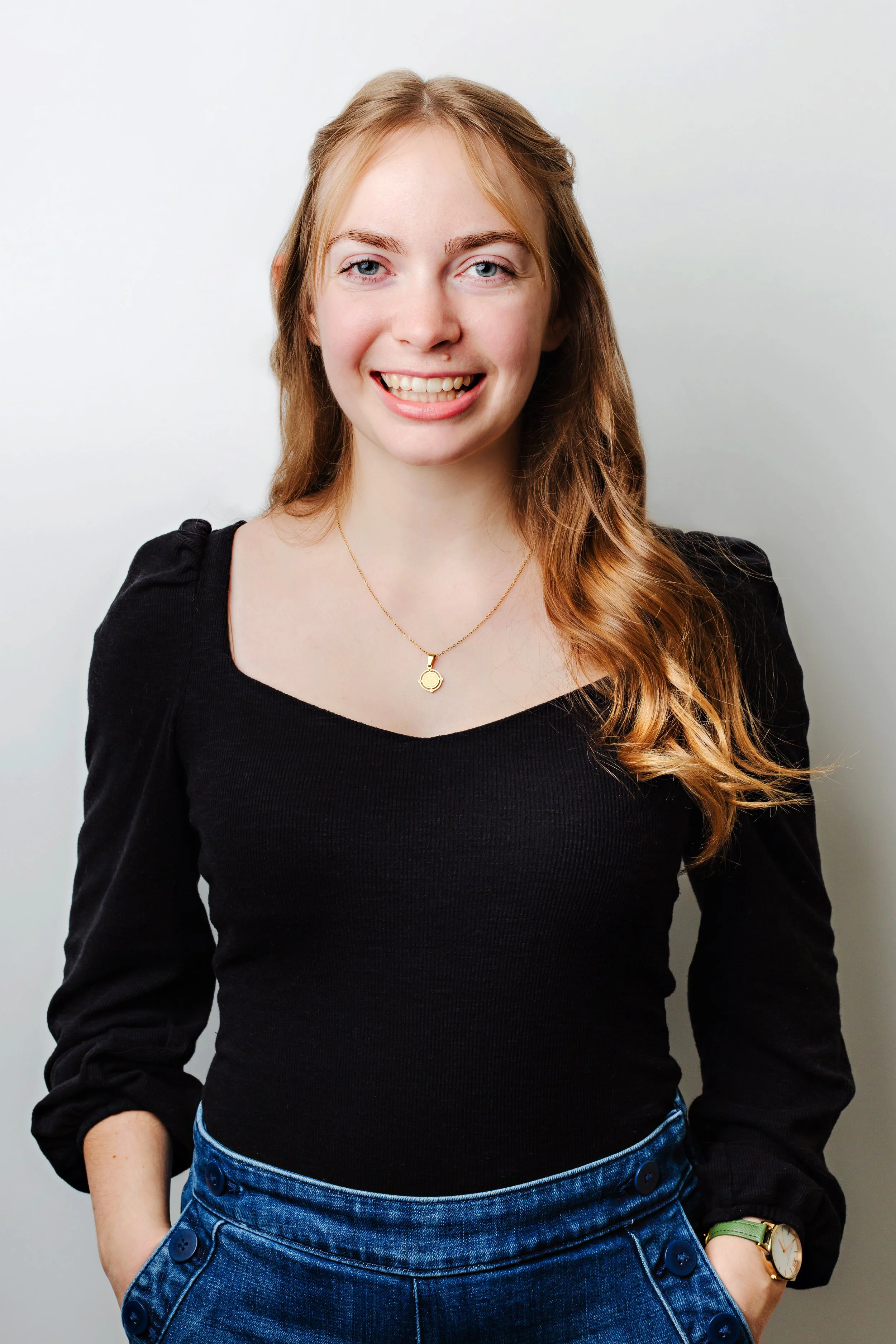 Qualifying Psychotherapist Emilie Chase — a young woman with long, wavy, reddish-blonde hair, smiling and posing against a gray background. She is wearing a black top, blue high-waisted jeans, and a gold watch.