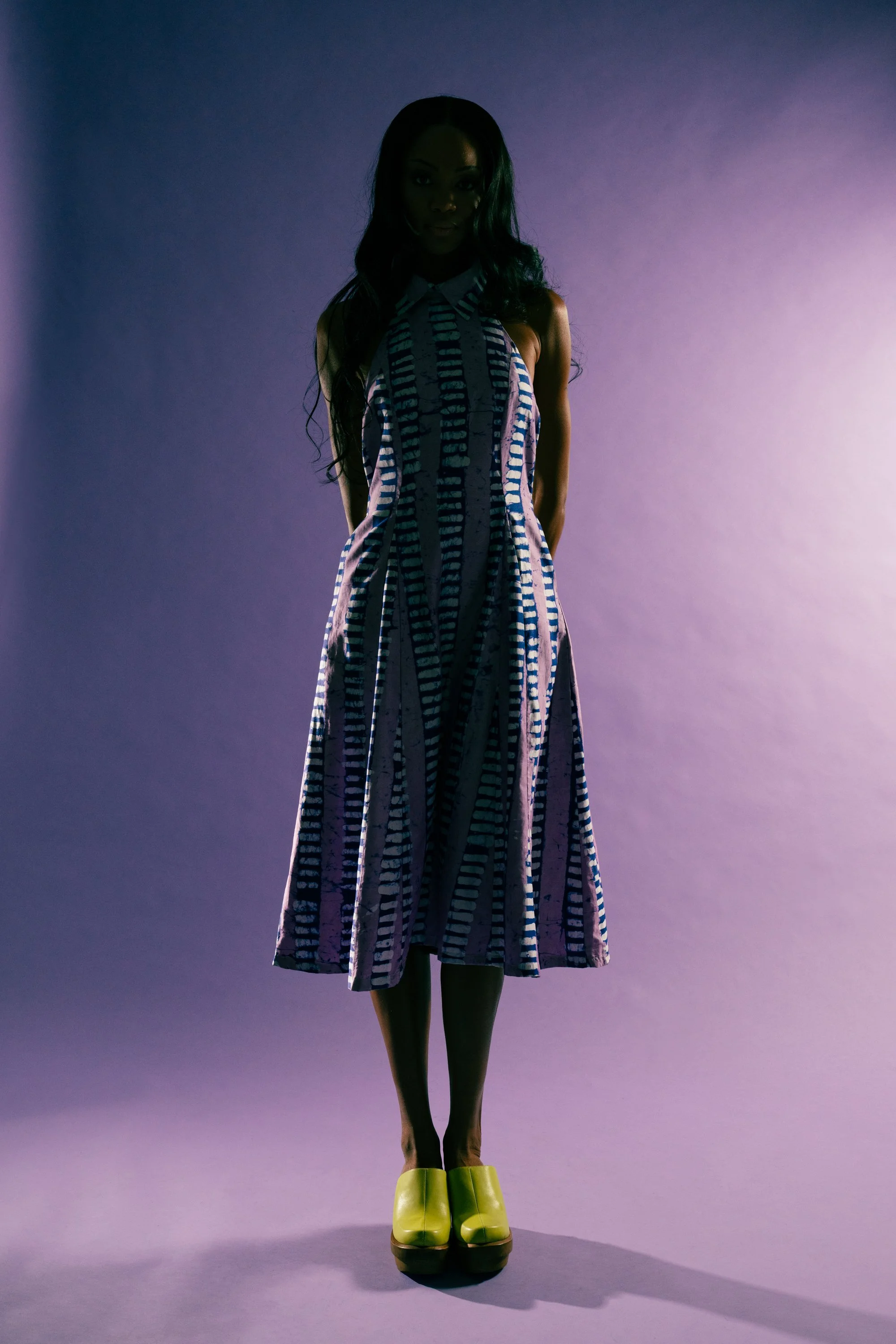 A woman in a colorful striped midi dress and yellow platform shoes standing against a purple backdrop.