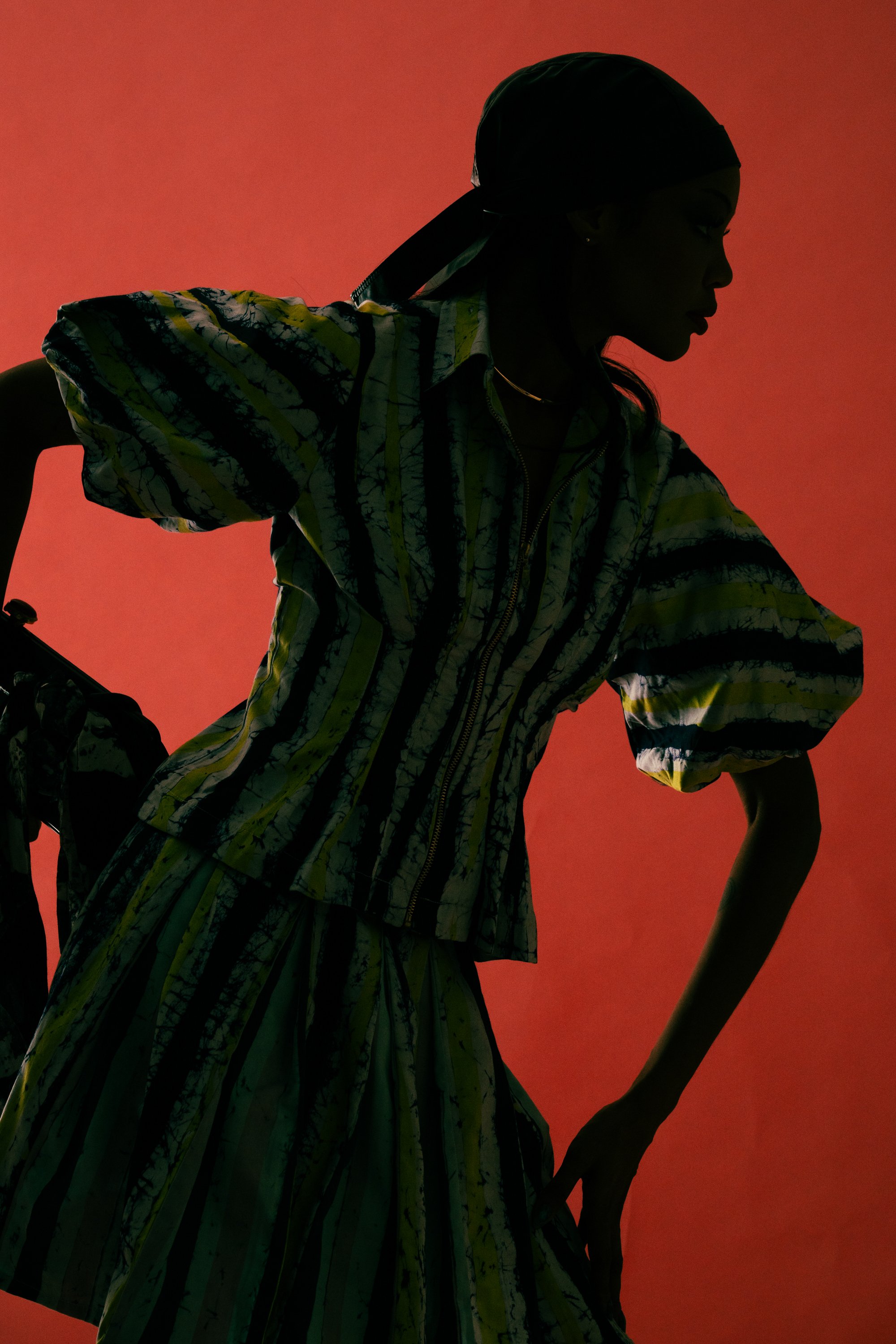 Silhouette of a woman in a patterned dress and headscarf, with her hands on her hips, against a red background.