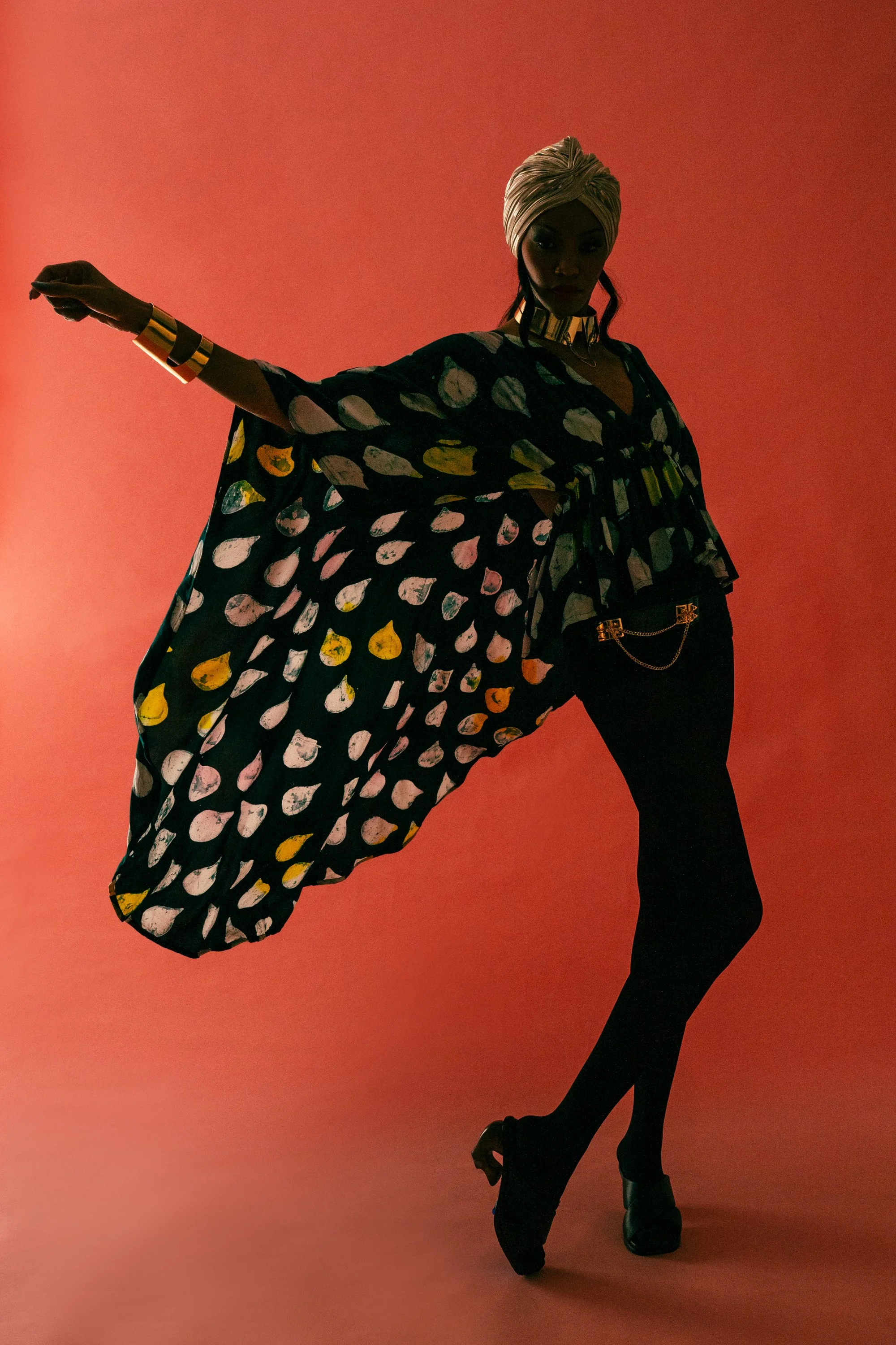 Fashion model striking a pose against a pink background, wearing patterned flowing cape, black pants, high heels, and accessorized with bold jewelry and a turban.