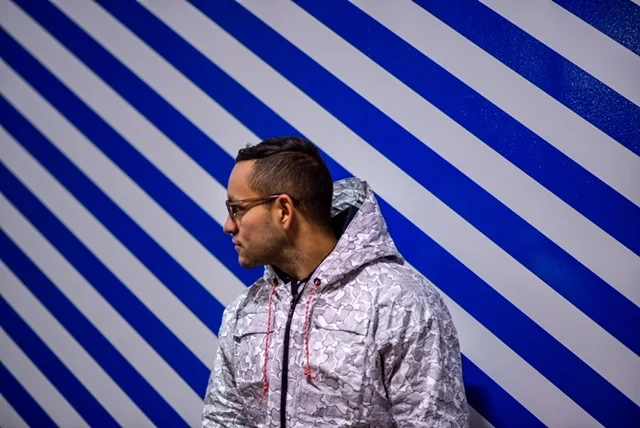a man against a blue and white striped wall wears a black and white jacket with a hood