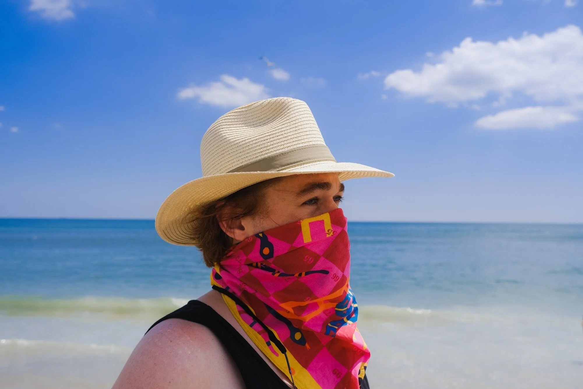 a man at the beach in a straw hat wears a bandana around his face like a mask