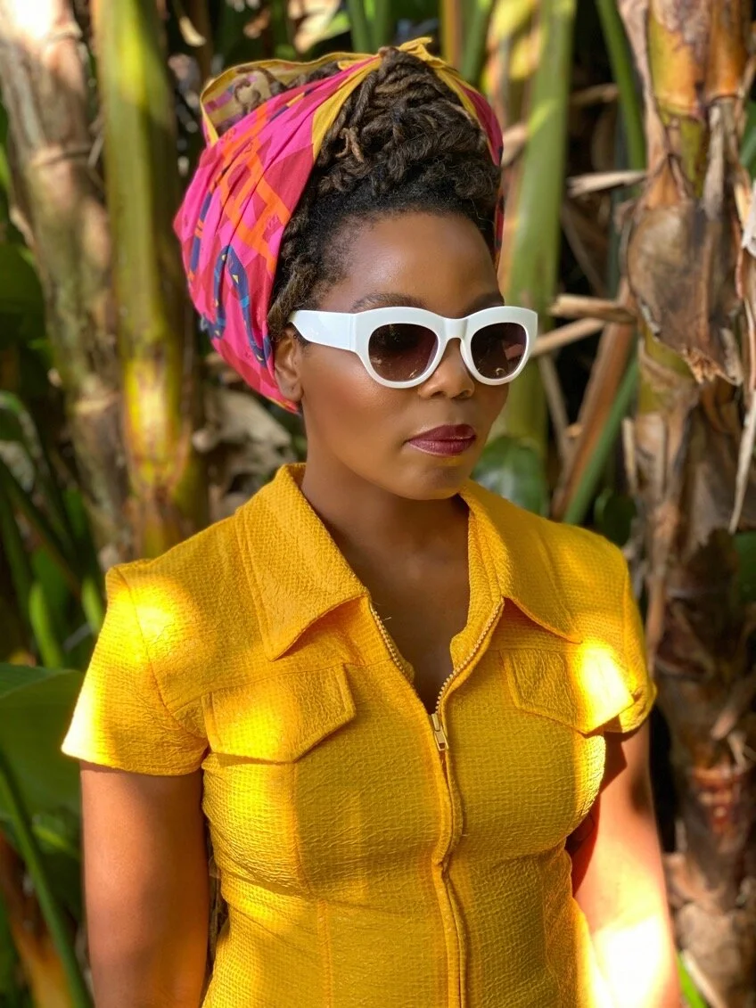 a woman in a yellow shirt and white sunglasses ties her hair back with a bandana scarf