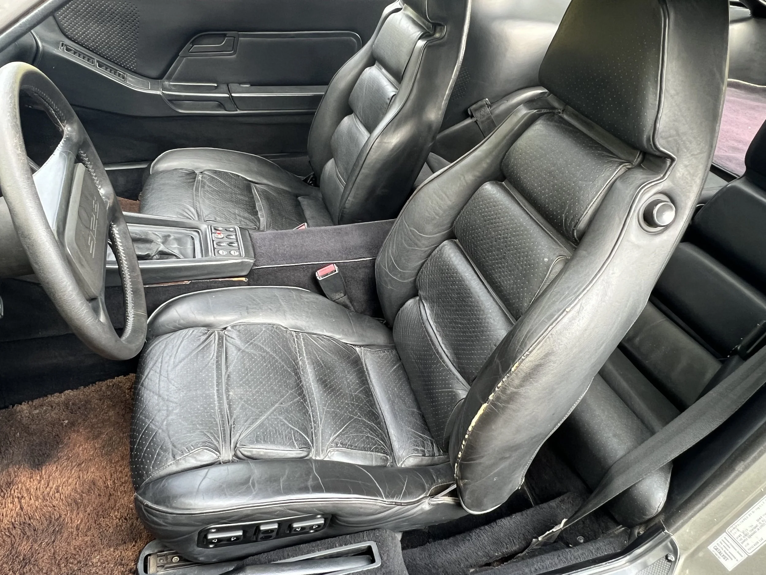Interior of a car showing black leather seats, some with visible wear and cracks, a brown carpeted floor, and a center console with controls.