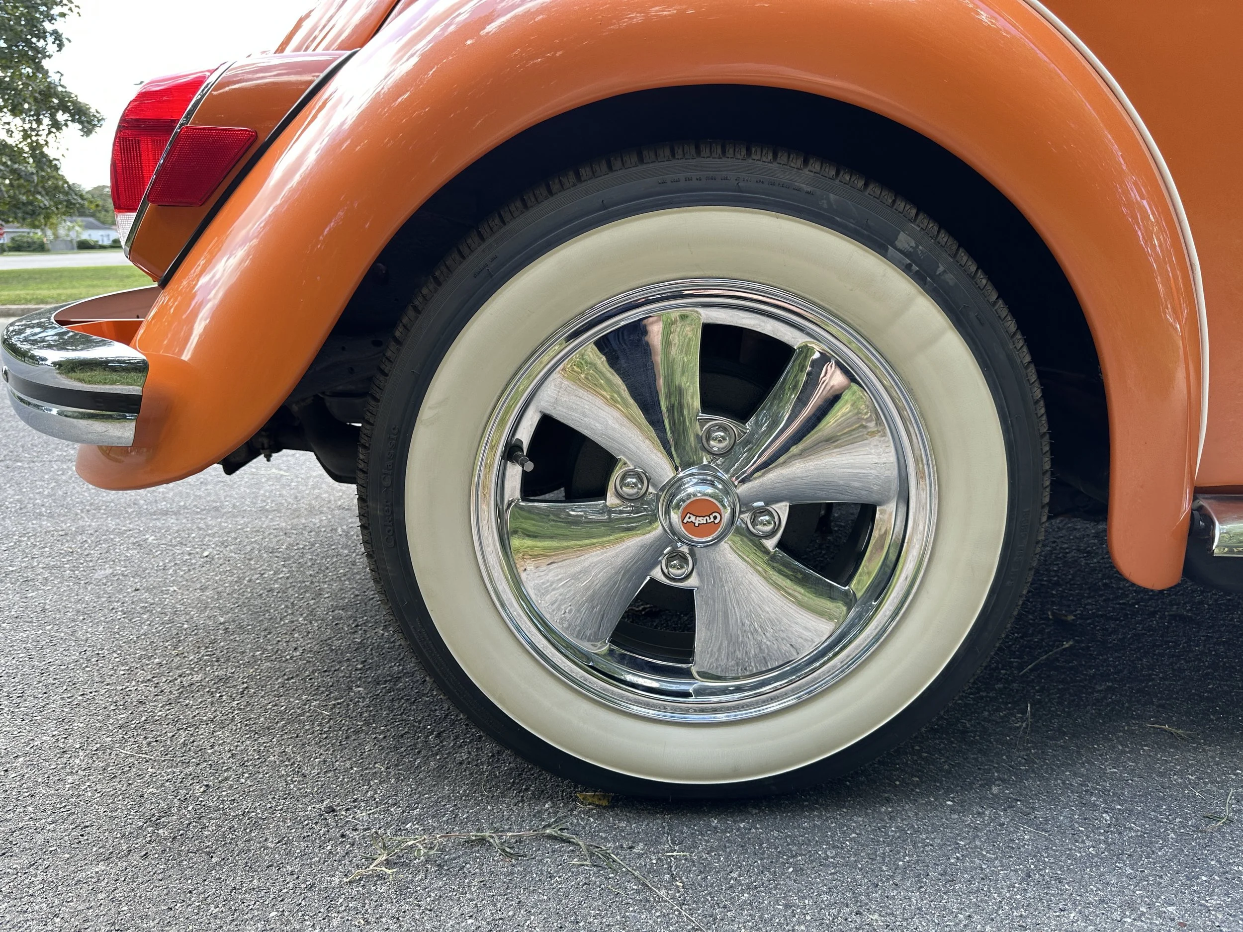Close-up of the front wheel and fender of an orange vintage car with cream-coloredwall tires and chrome hubcap.