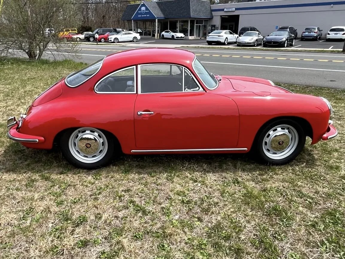 A red vintage Porsche 356 sports car parked on grass outside a car dealership with multiple modern cars in the background.