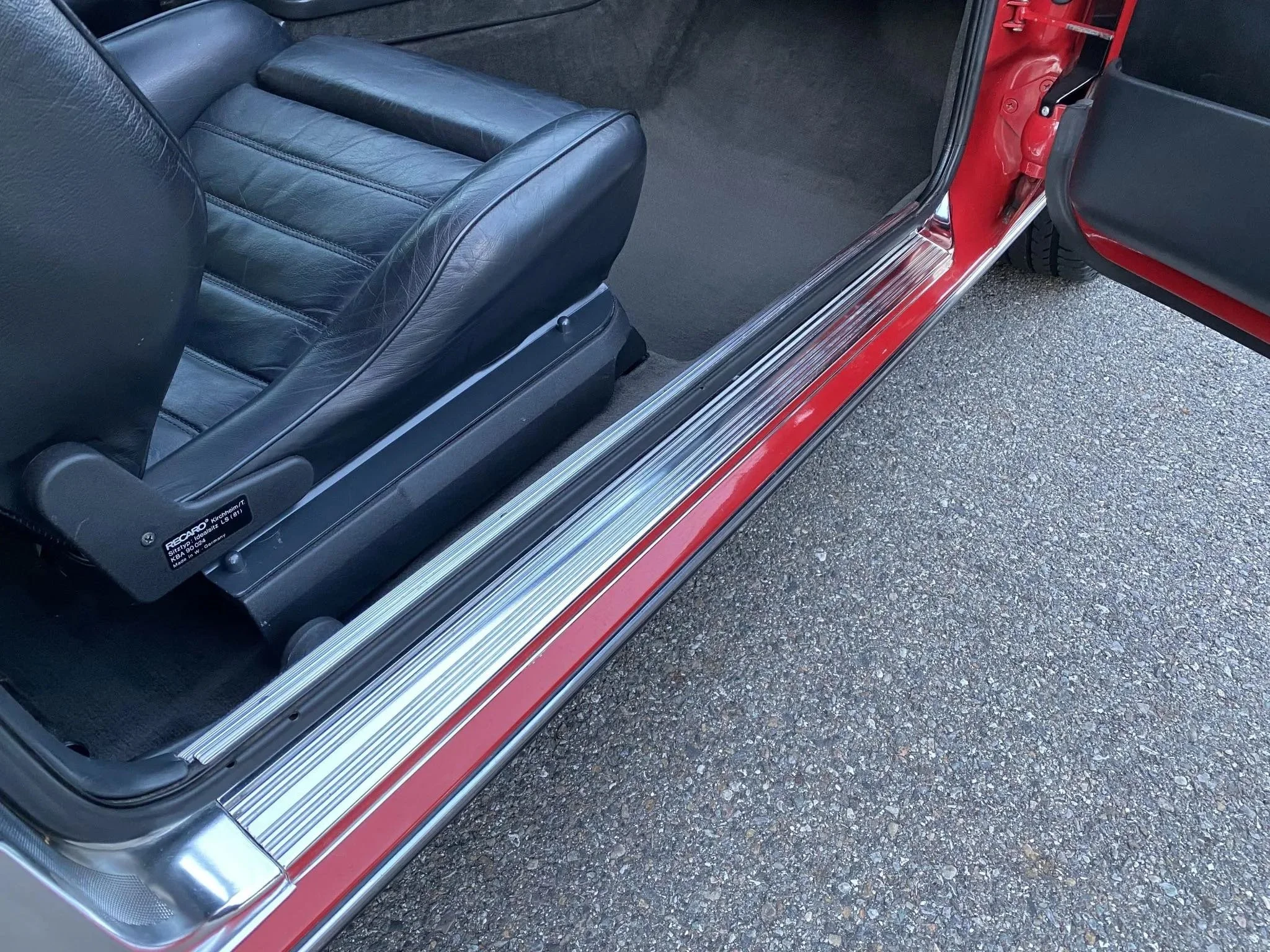 Close-up of the interior of a red vintage car, showing a black leather front seat and door sill with chrome trim.
