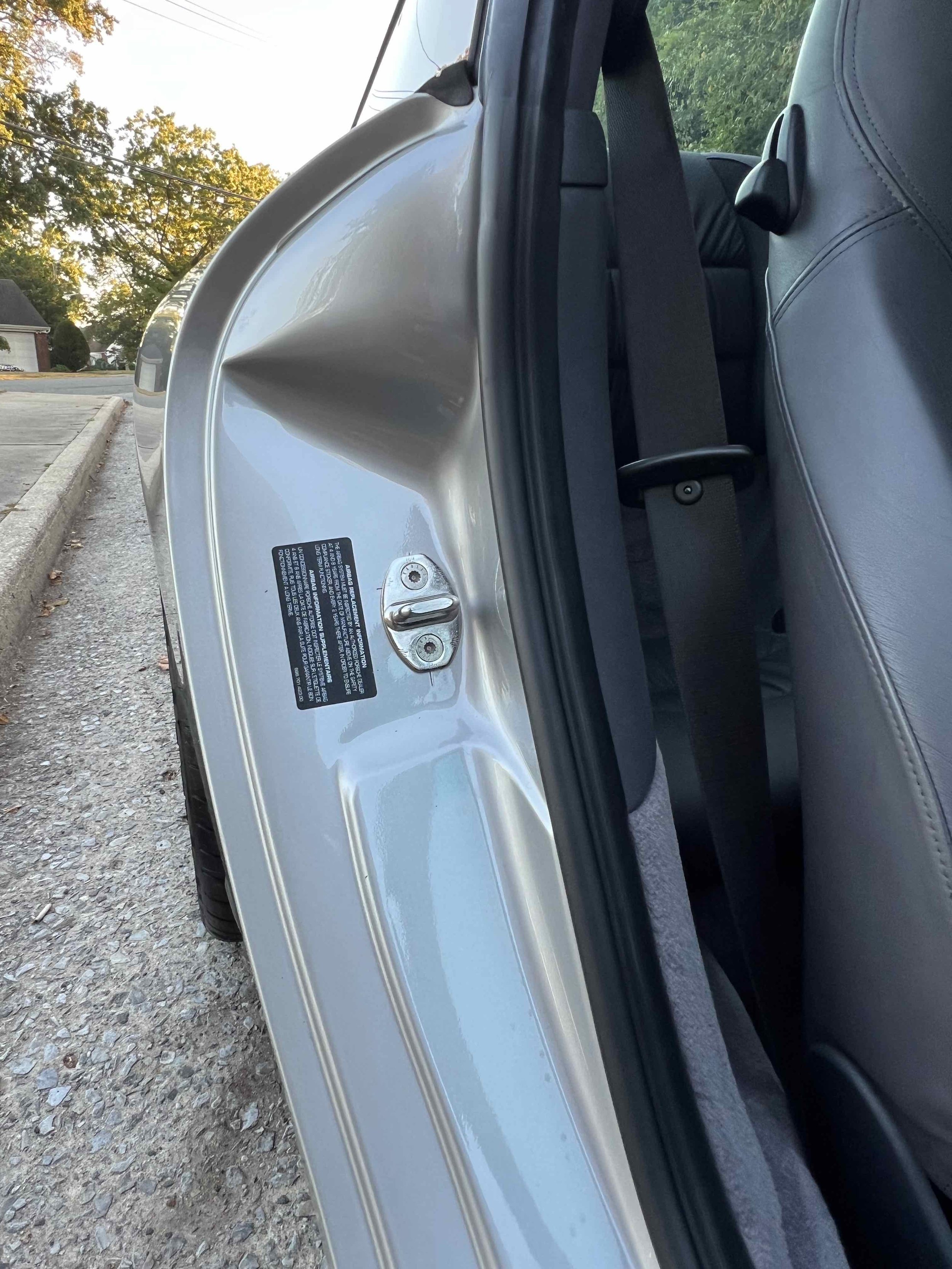 The photo shows the interior side of a car door, with the door open, revealing the latch mechanism and a black sticker with information. The exterior of the car is silver, and the interior features gray leather seats. The background includes a concre