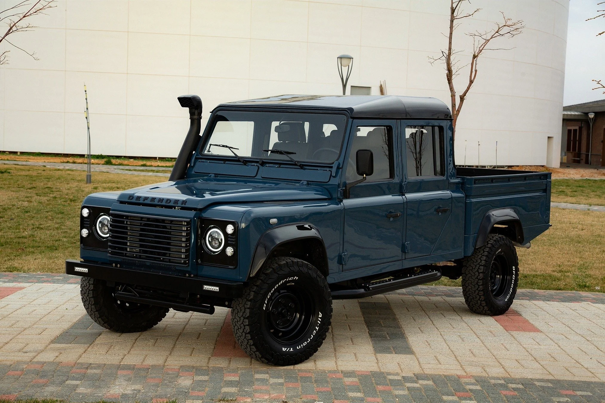 A dark blue Land Rover Defender pickup truck parked on a paved area with a grassy background and a modern building.