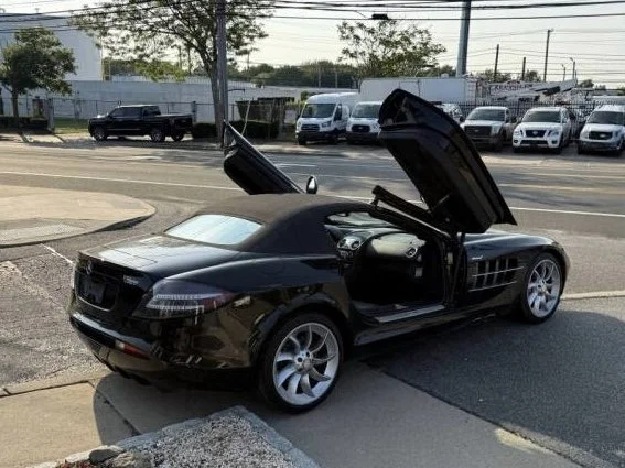 A small black toy car with gullwing doors open parked on the street with parked cars and trees in the background.
