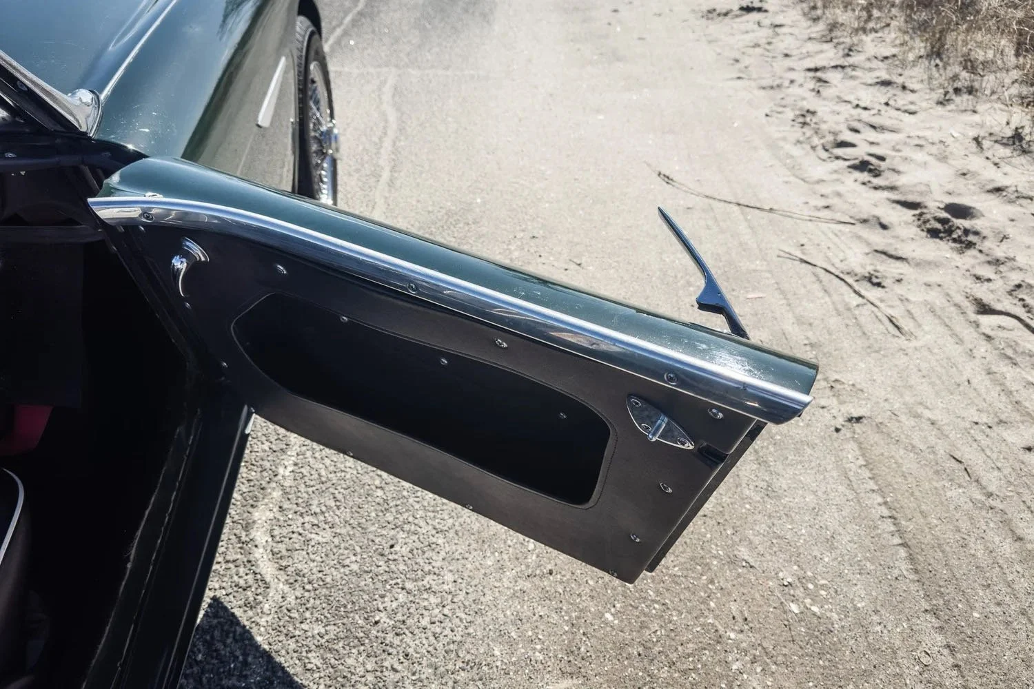 Close-up of an open vintage car door with a black interior and chrome trim, parked on a sandy road.