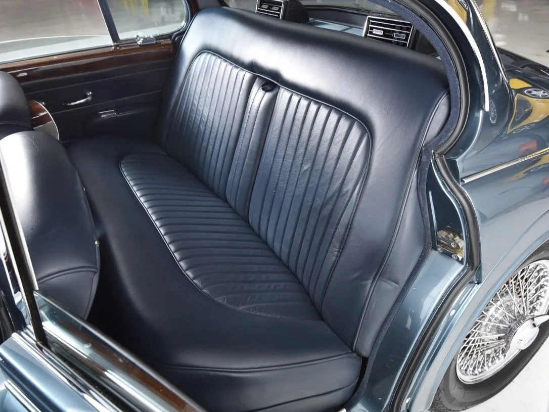 The image shows the interior of a vintage car, featuring a black leather back seat with vertical stitching, wood paneling on the door, and a wire-spoked wheel visible on the exterior.