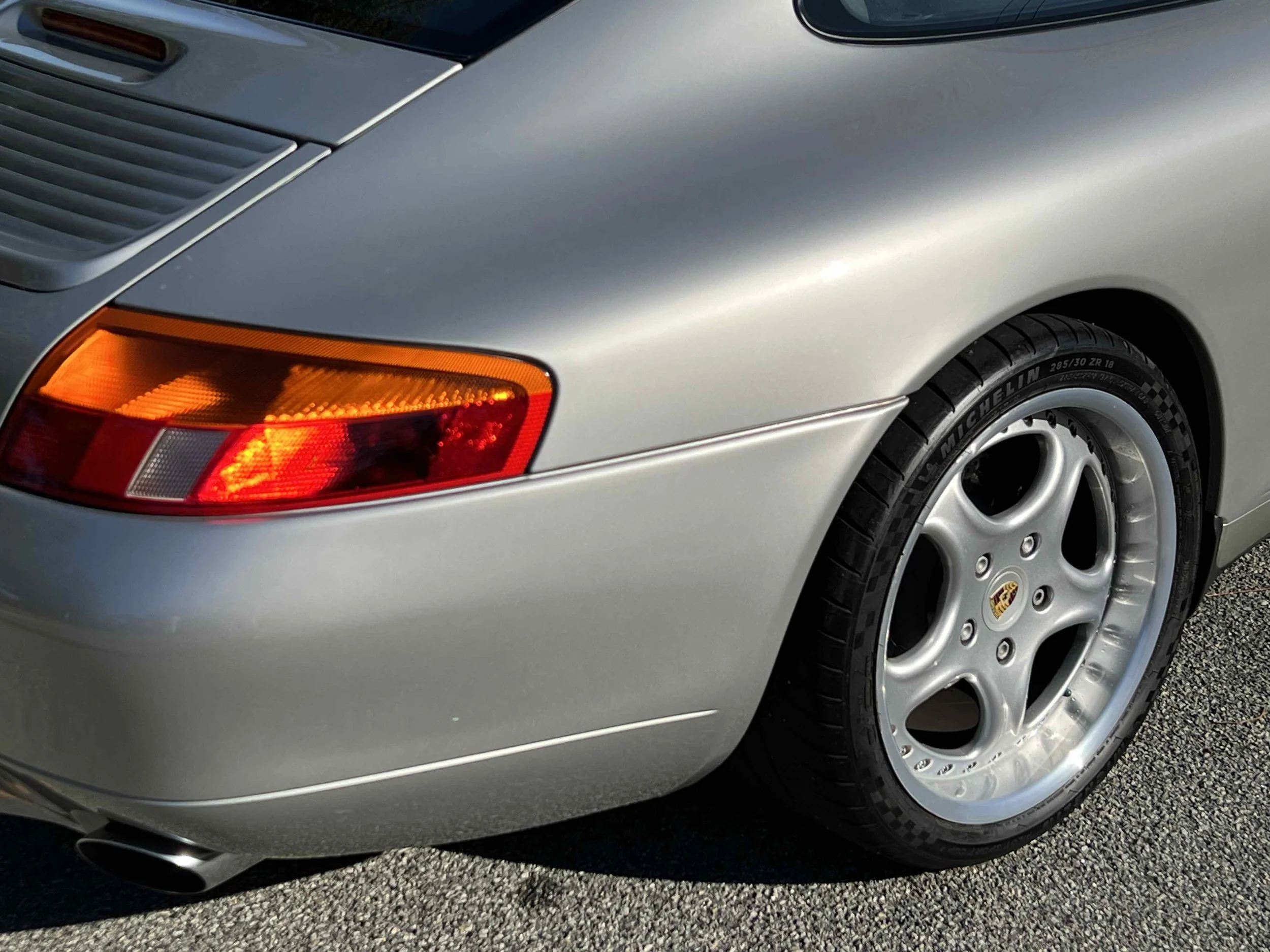 Close-up of the rear side of a silver Porsche 911, showing the tail light, part of the rear wheel and tire, and the exhaust pipe.