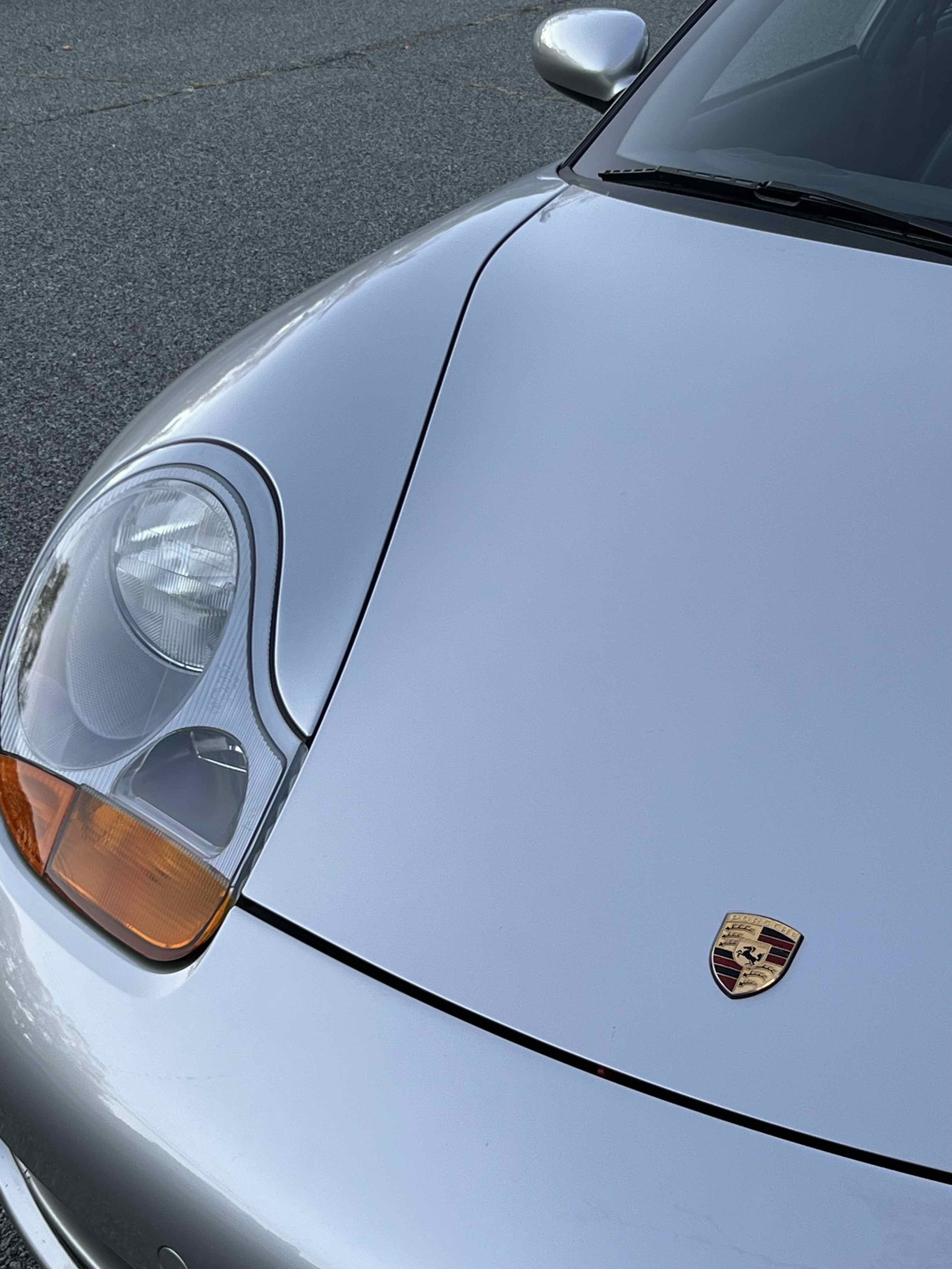 Close-up of silver Porsche car, showing front headlight, part of the hood with Porsche emblem, and side mirror on the driver's side