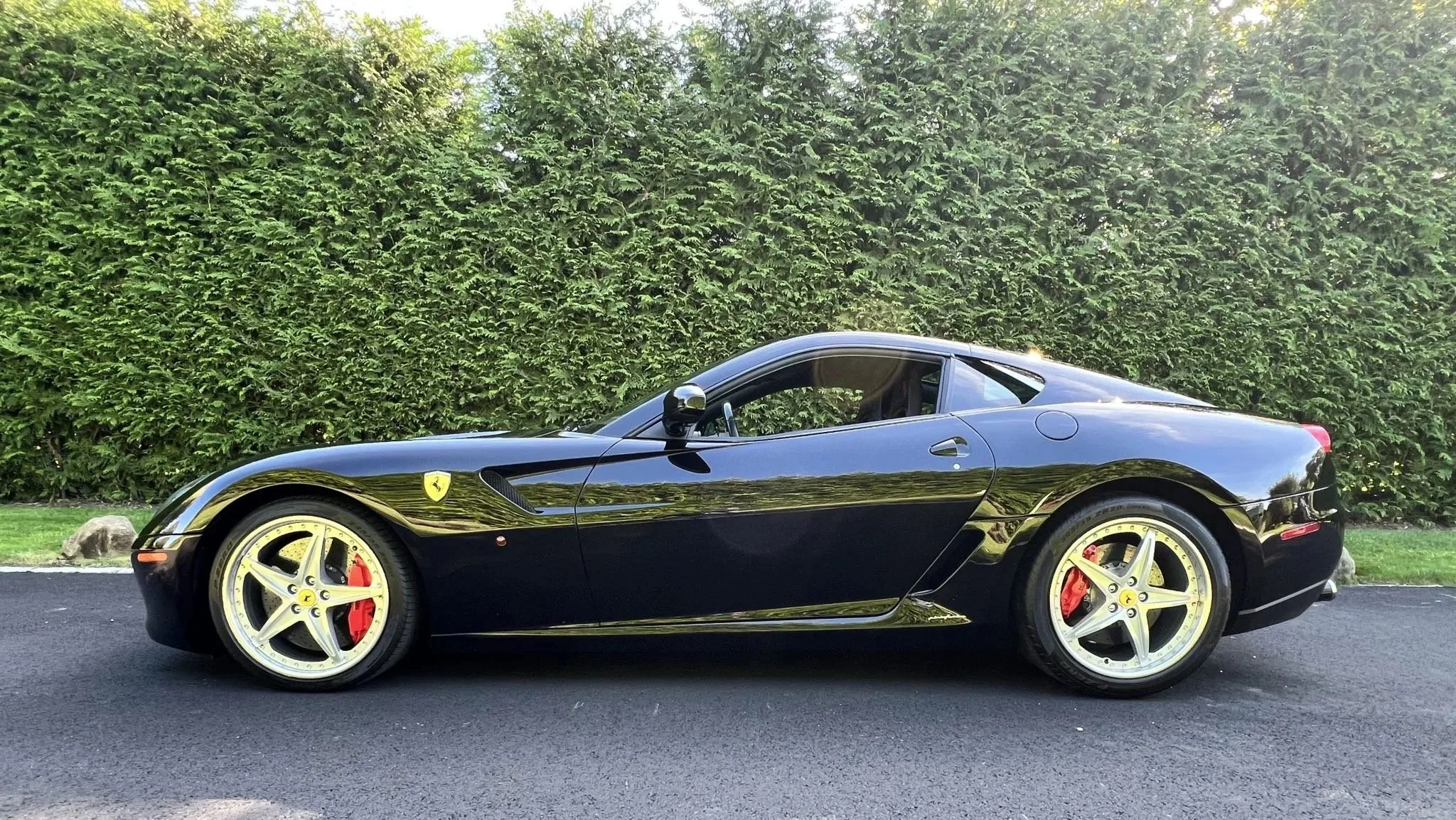 A black Ferrari sports car parked on the road with a lush green hedge in the background.