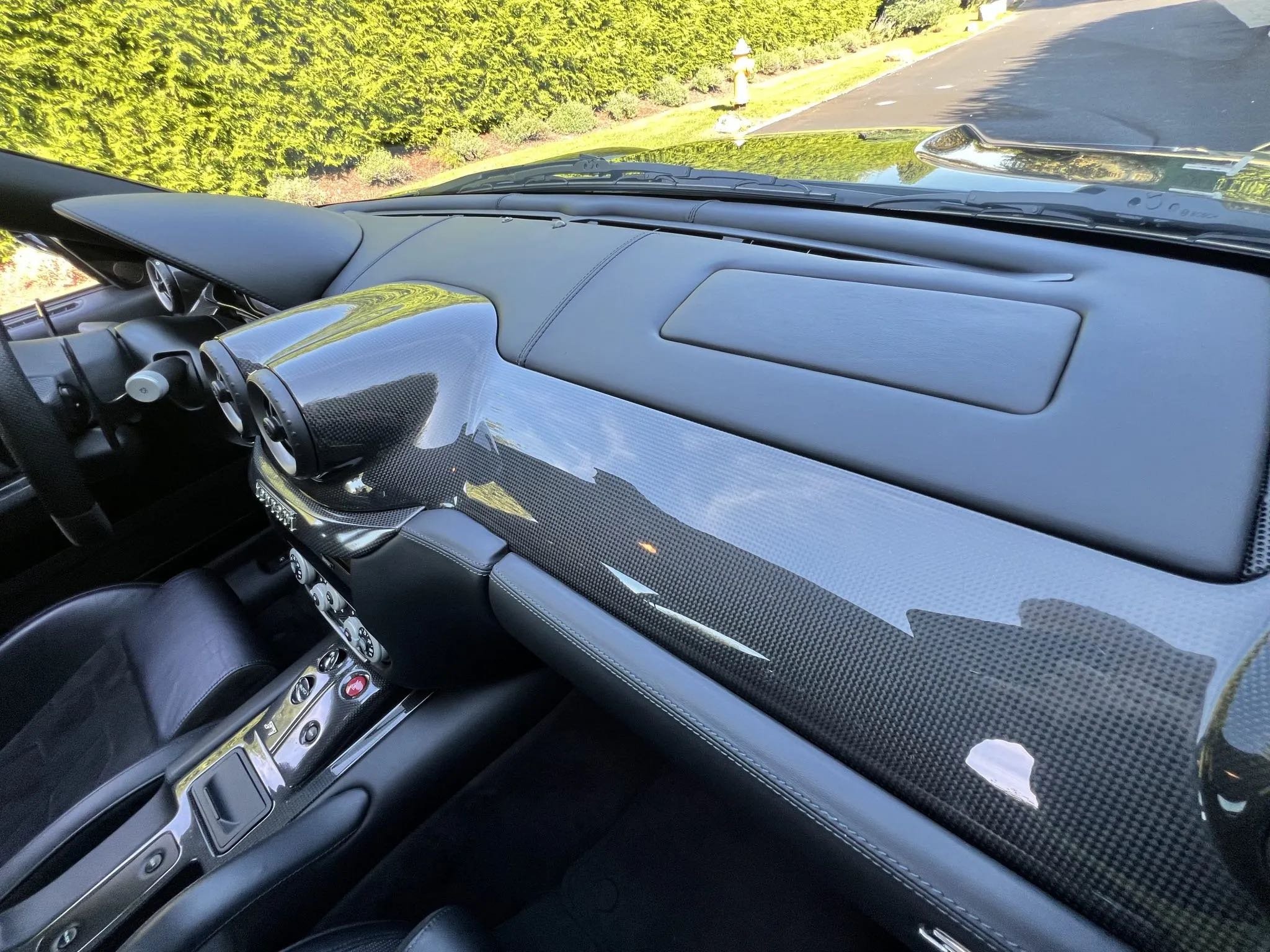 The interior of a luxury sports car showing a carbon fiber dashboard, air vents, and part of the steering wheel, with a view of the outside road and greenery through the windshield.