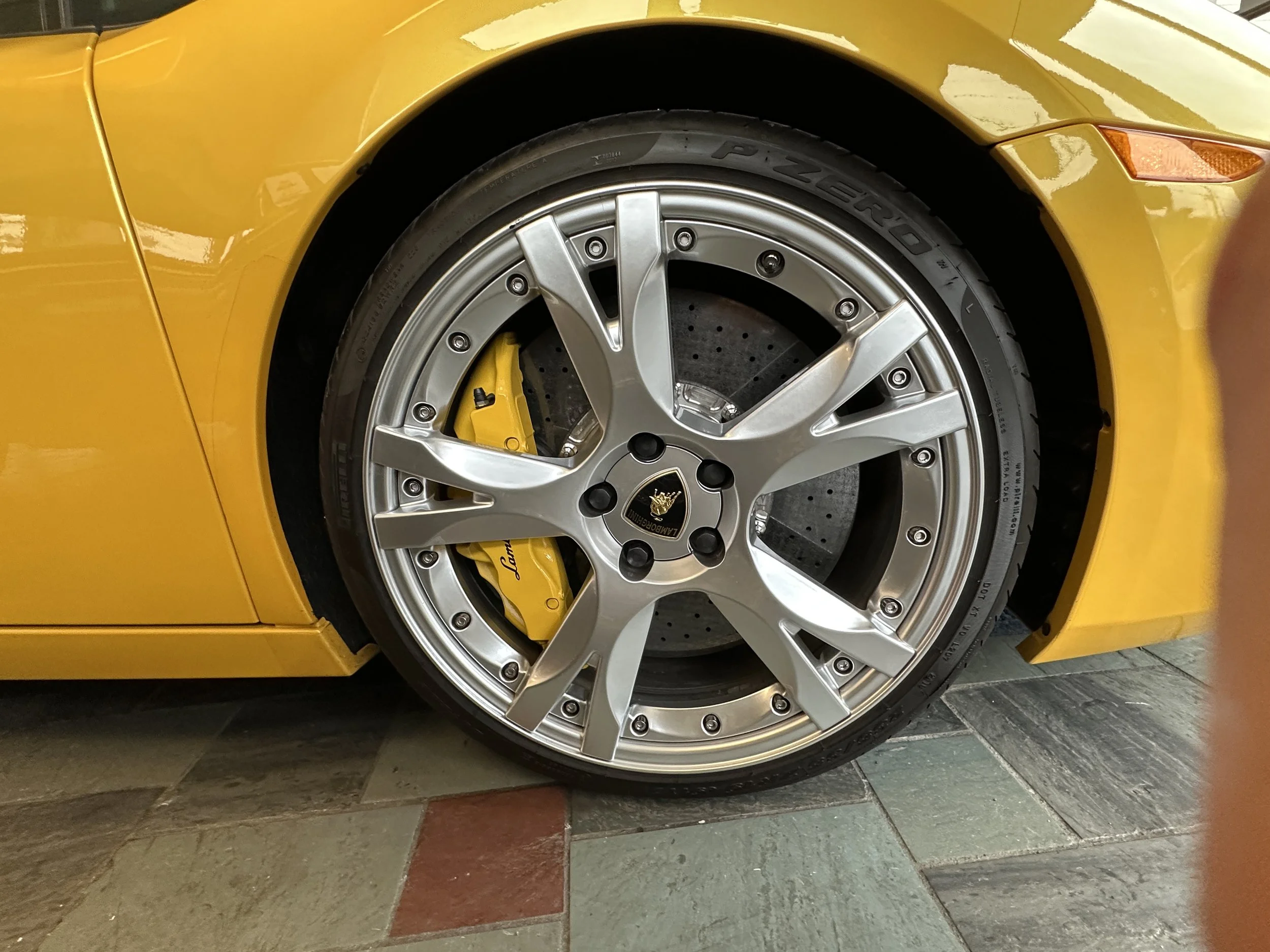 Close-up of a yellow sports car's front wheel showing a silver alloy rim, yellow brake caliper, and drilled brake disc.
