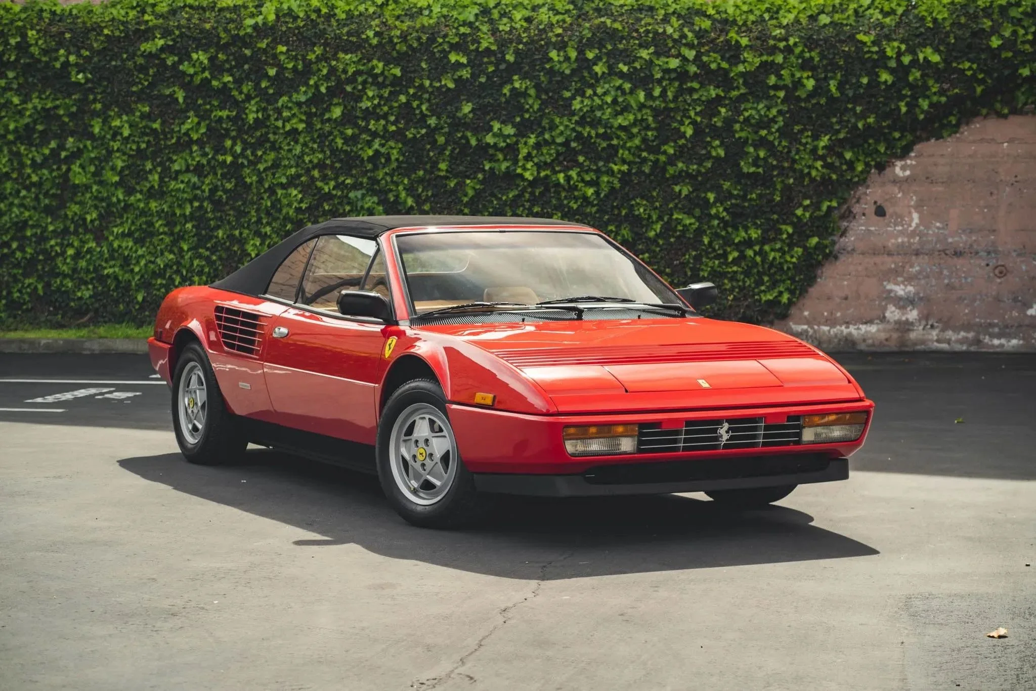 Red vintage Ferrari sports car parked on asphalt with green bushes in background.
