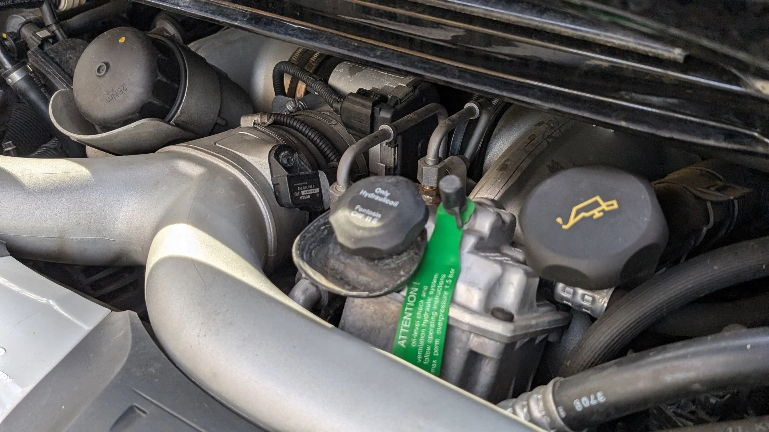Close-up of a car engine showing fluid caps, hoses, and components including a green warning tag.