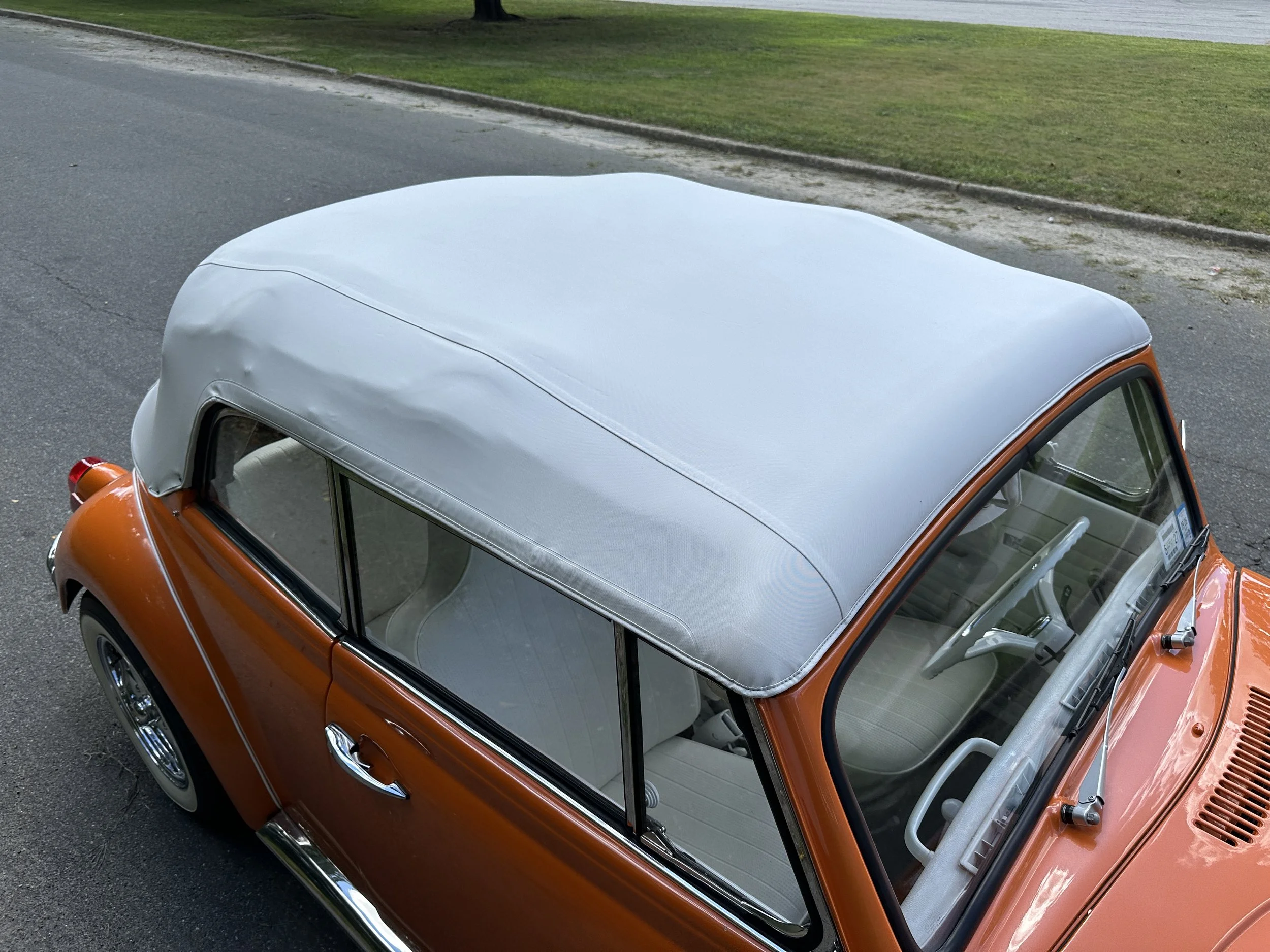 Top view of a vintage orange convertible car with a light gray soft top, parked on a paved street near a grassy area.