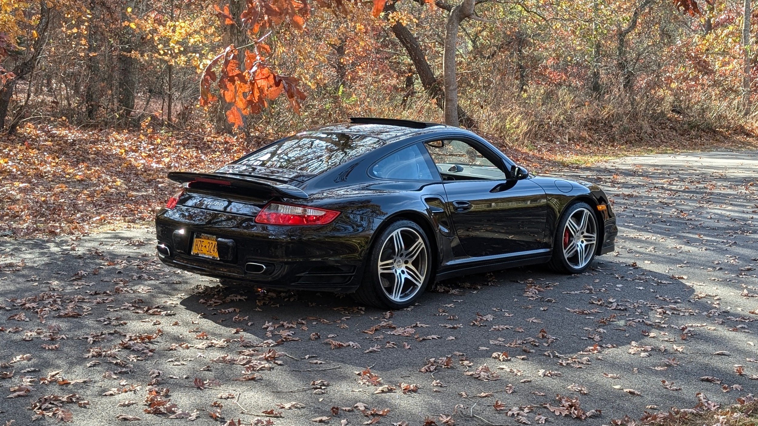 Black Porsche sports car parked on a leaf-covered asphalt road in a wooded area during fall, with trees showing autumn foliage in the background.