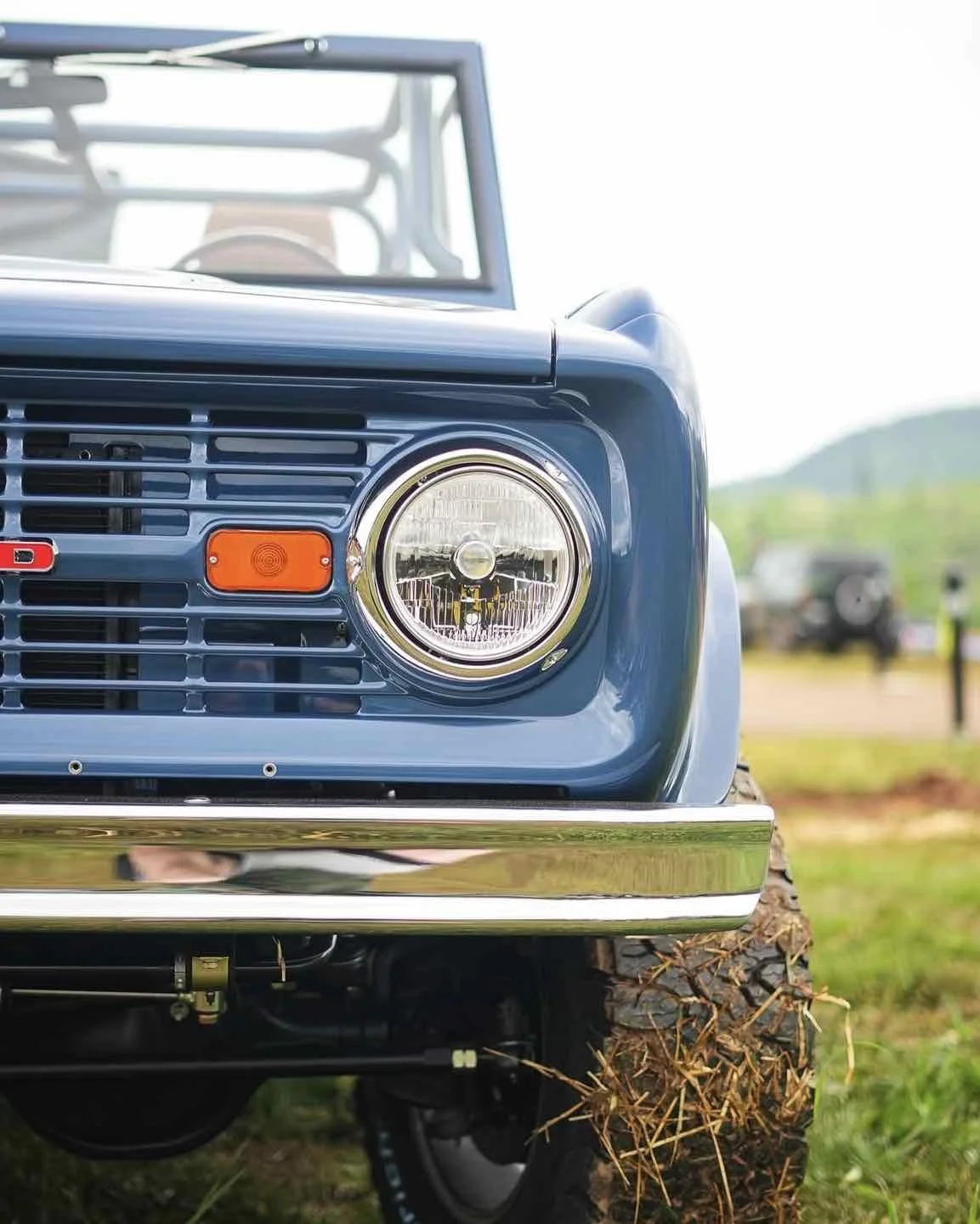 Close-up of the front of a vintage blue off-road vehicle showing the right headlight, grille, and part of the bumper, with a grassy field and other vehicles in the background.
