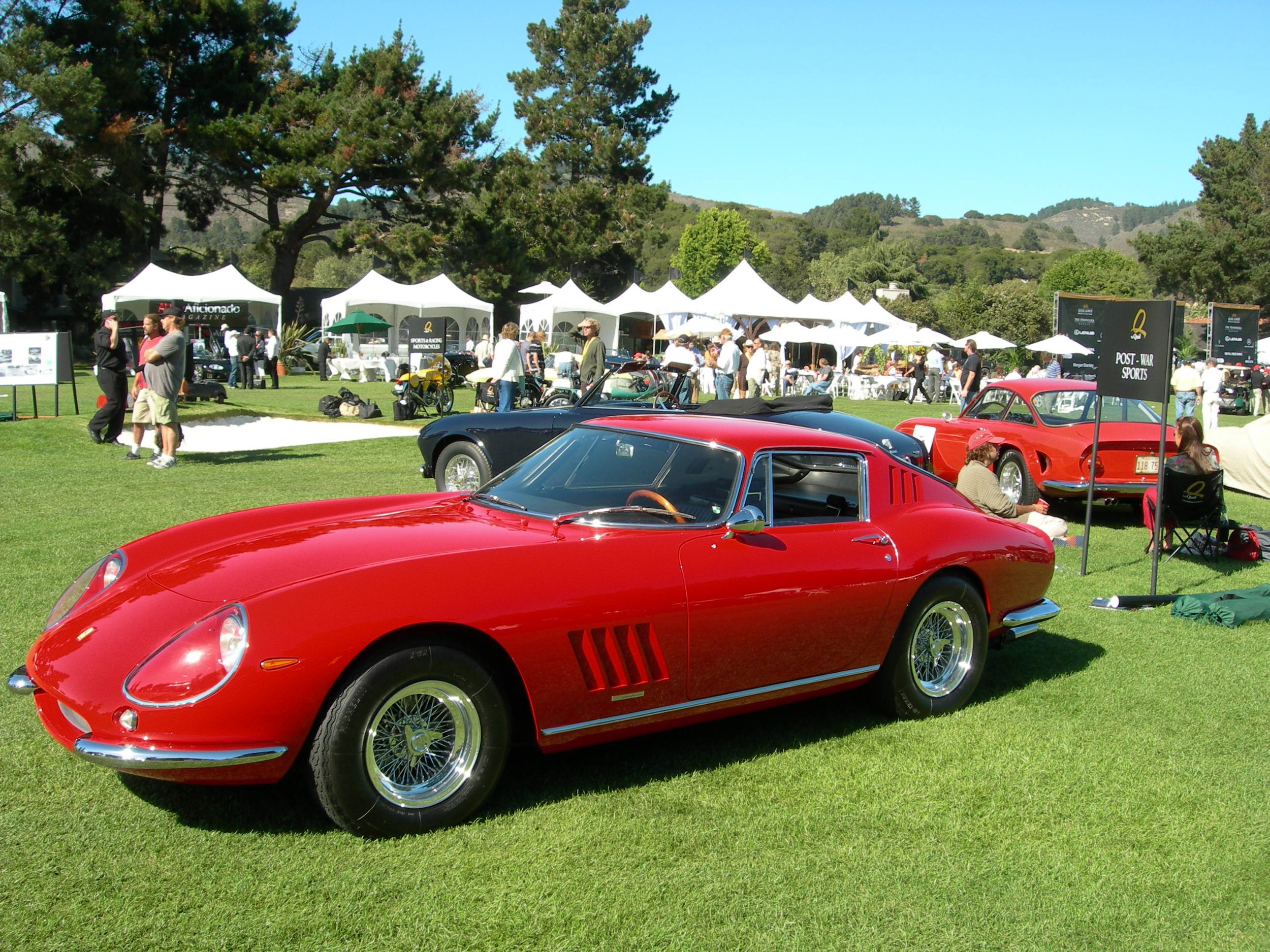 Red vintage sports car at a classic car show on a grassy field with white tents and people in the background.